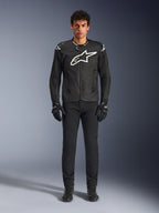 Alpinestars T-GP Air Jacket, zwarte textielen motorjas met een groot wit Astars-logo op de borst, gedragen door een model met zwarte motorbroek en handschoenen, voorzien van geperforeerde panelen en mesh-ventilatie voor sportief rijden