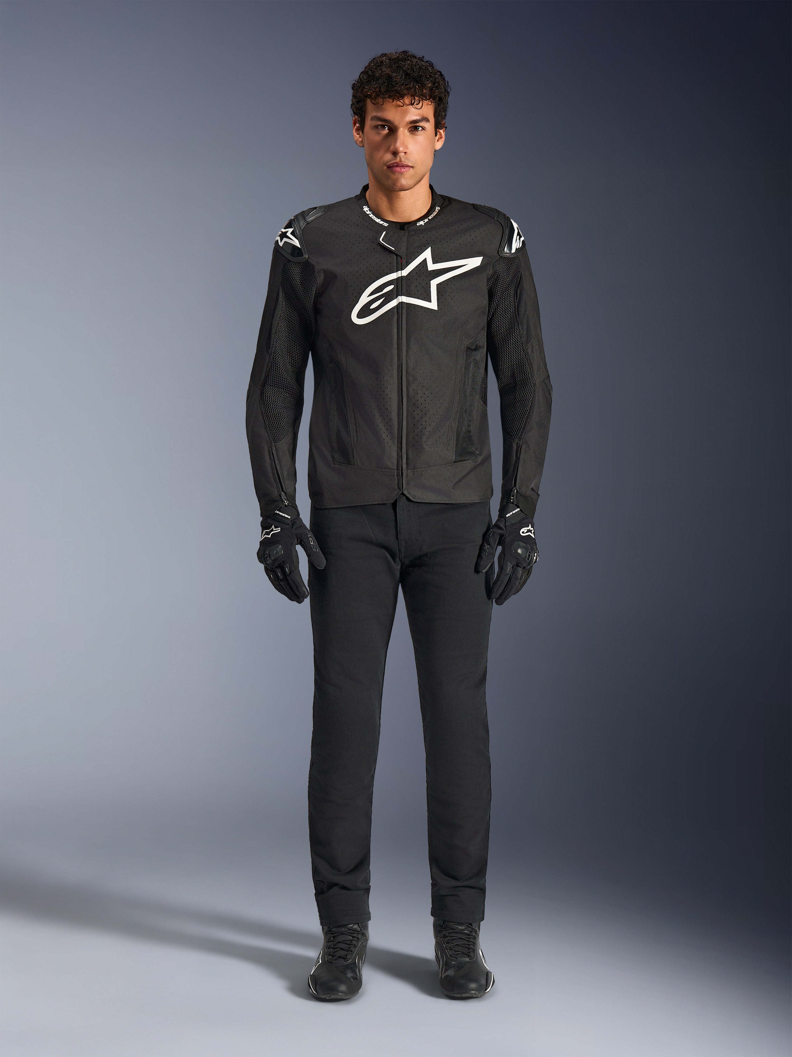 Alpinestars T-GP Air Jacket, zwarte textielen motorjas met een groot wit Astars-logo op de borst, gedragen door een model met zwarte motorbroek en handschoenen, voorzien van geperforeerde panelen en mesh-ventilatie voor sportief rijden