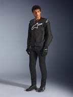 Alpinestars T-GP Air Jas, zwarte textiel motorjas, gedragen door een model met bijpassende zwarte broek en handschoenen, met een opvallend wit Alpinestars-logo op de borst en schouders, geventileerde mesh-panelen en een sportieve, van de racerij afgeleide pasvorm