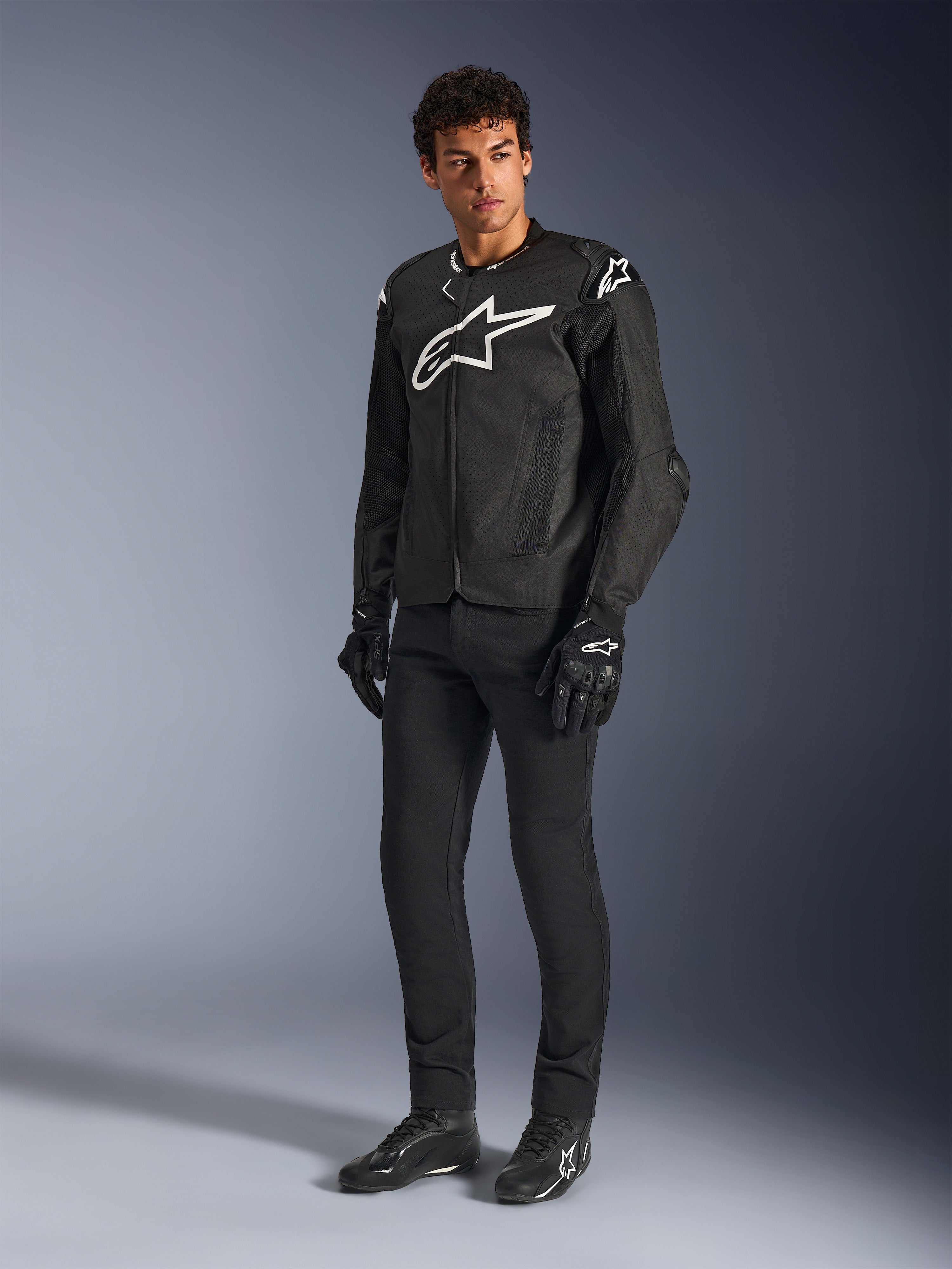 Alpinestars T-GP Air Jas, zwarte textiel motorjas, gedragen door een model met bijpassende zwarte broek en handschoenen, met een opvallend wit Alpinestars-logo op de borst en schouders, geventileerde mesh-panelen en een sportieve, van de racerij afgeleide pasvorm
