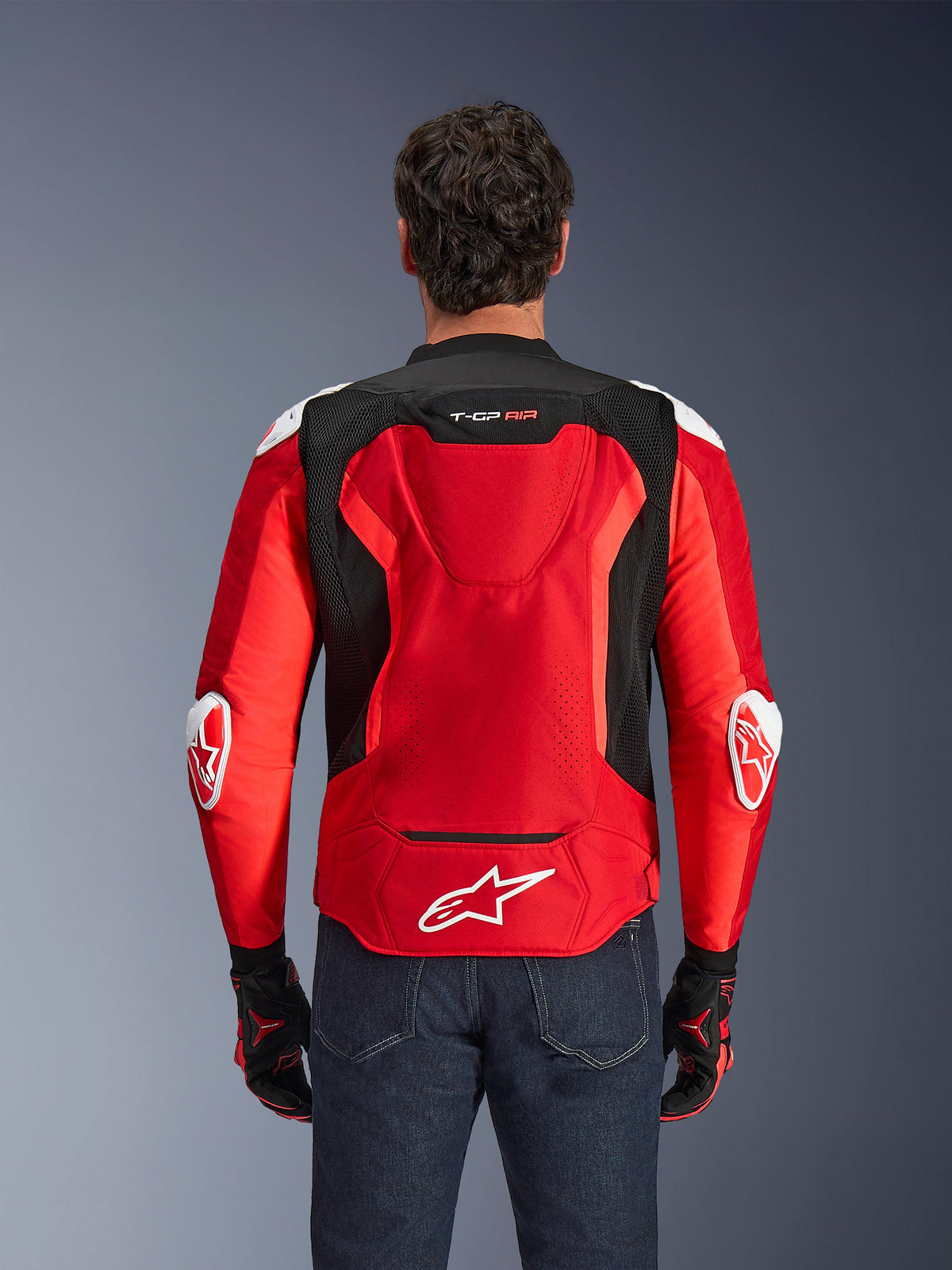 Alpinestars T-GP Air Jacket, textiel motorjas, felrood en zwart met witte accenten, achteraanzicht met de aero-bult en mesh-ventilatiepanelen, voorzien van het Alpinestars-sterlogo en T-GP Air-branding, gedragen met een donkere jeans en racehandschoenen