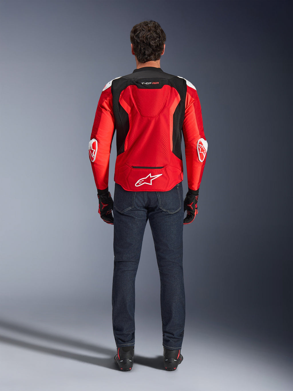 Alpinestars T-GP Air motorjas, achteraanzicht, felrood en zwart, met een wit logo op de onderrug en 'T-GP AIR'-branding, gedragen met een donkerblauwe spijkerbroek en zwart-rode motorhandschoenen, sportieve motoruitrusting