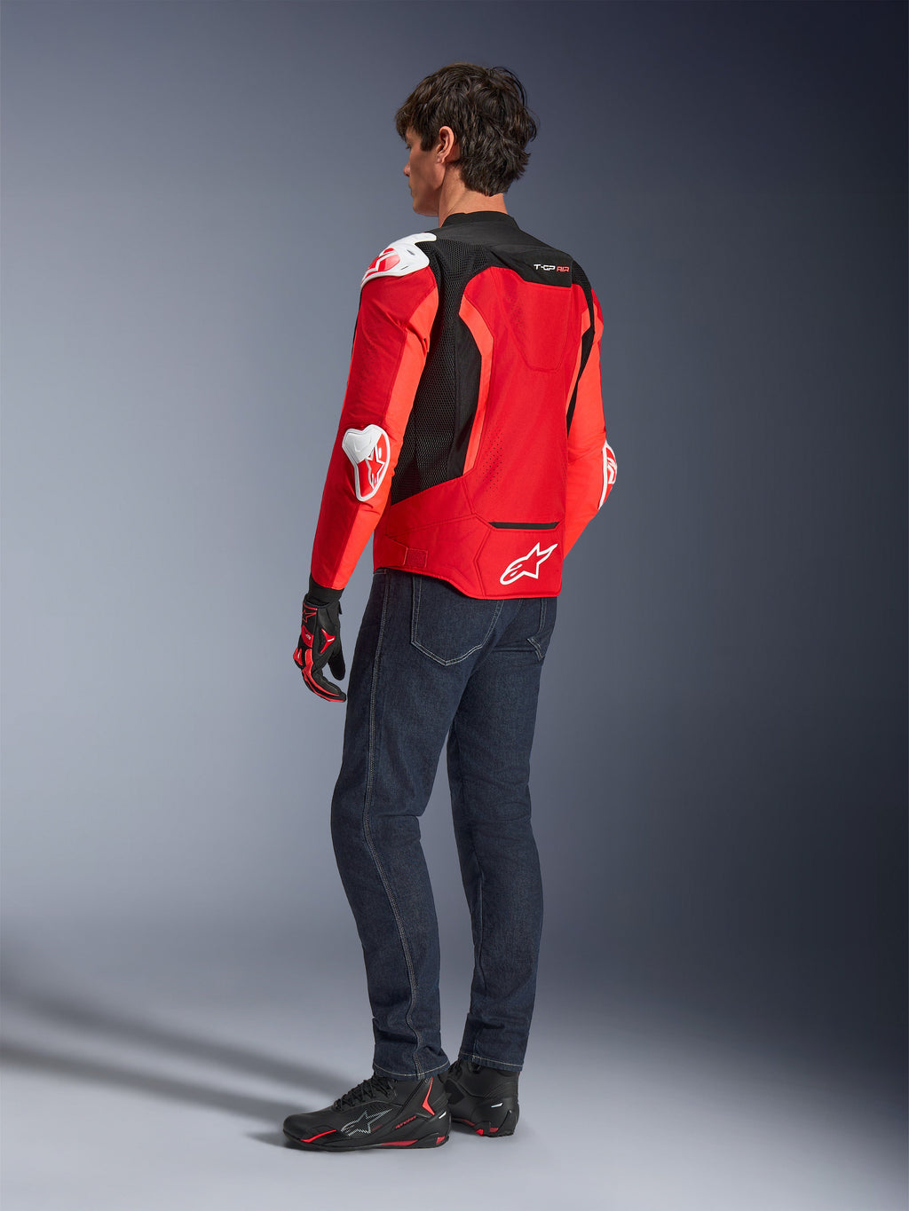 Alpinestars T-GP Air Jacket, achteraanzicht van een man die een felrode en fluo rode textieljas draagt met zwarte mesh-panelen en witte sliders, gecombineerd met een donkerblauwe jeans en zwarte motorschoenen, sportieve motoruitrusting
