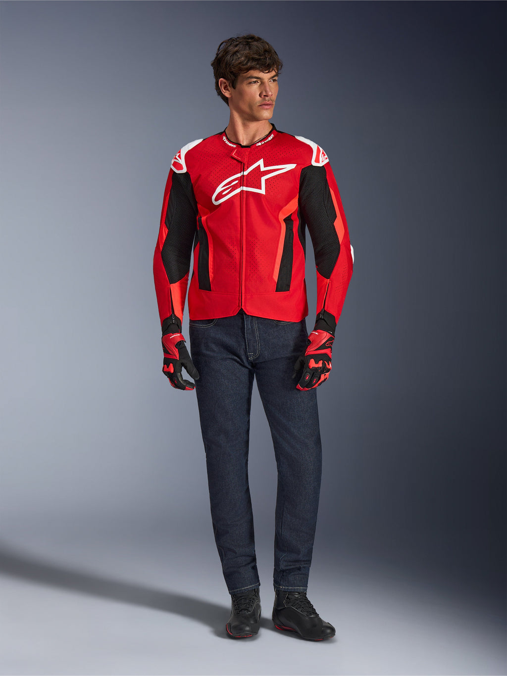 Alpinestars T-GP Air Jacket, helderrood fluorrood zwart, mannelijk model in volledige motoroutfit met donkerblauwe jeans en bijpassende handschoenen, sportieve textieljas met geperforeerde mesh-panelen en wit Astars-logo, studiofoto.