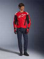 Alpinestars T-GP Air Jacket, helderrood fluorrood zwart, mannelijk model in volledige motoroutfit met donkerblauwe jeans en bijpassende handschoenen, sportieve textieljas met geperforeerde mesh-panelen en wit Astars-logo, studiofoto.