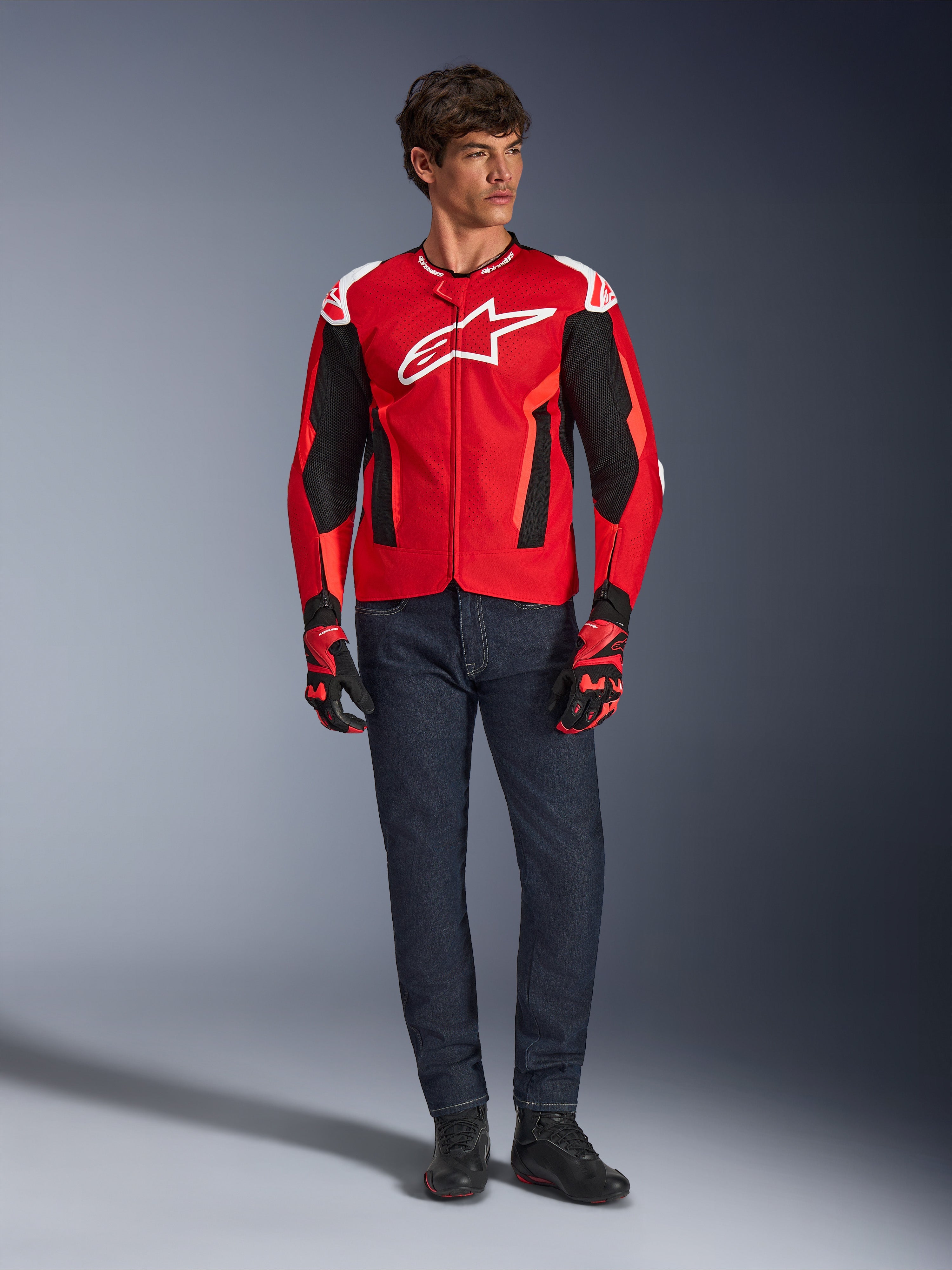Alpinestars T-GP Air Jacket, helderrood fluorrood zwart, mannelijk model in volledige motoroutfit met donkerblauwe jeans en bijpassende handschoenen, sportieve textieljas met geperforeerde mesh-panelen en wit Astars-logo, studiofoto.