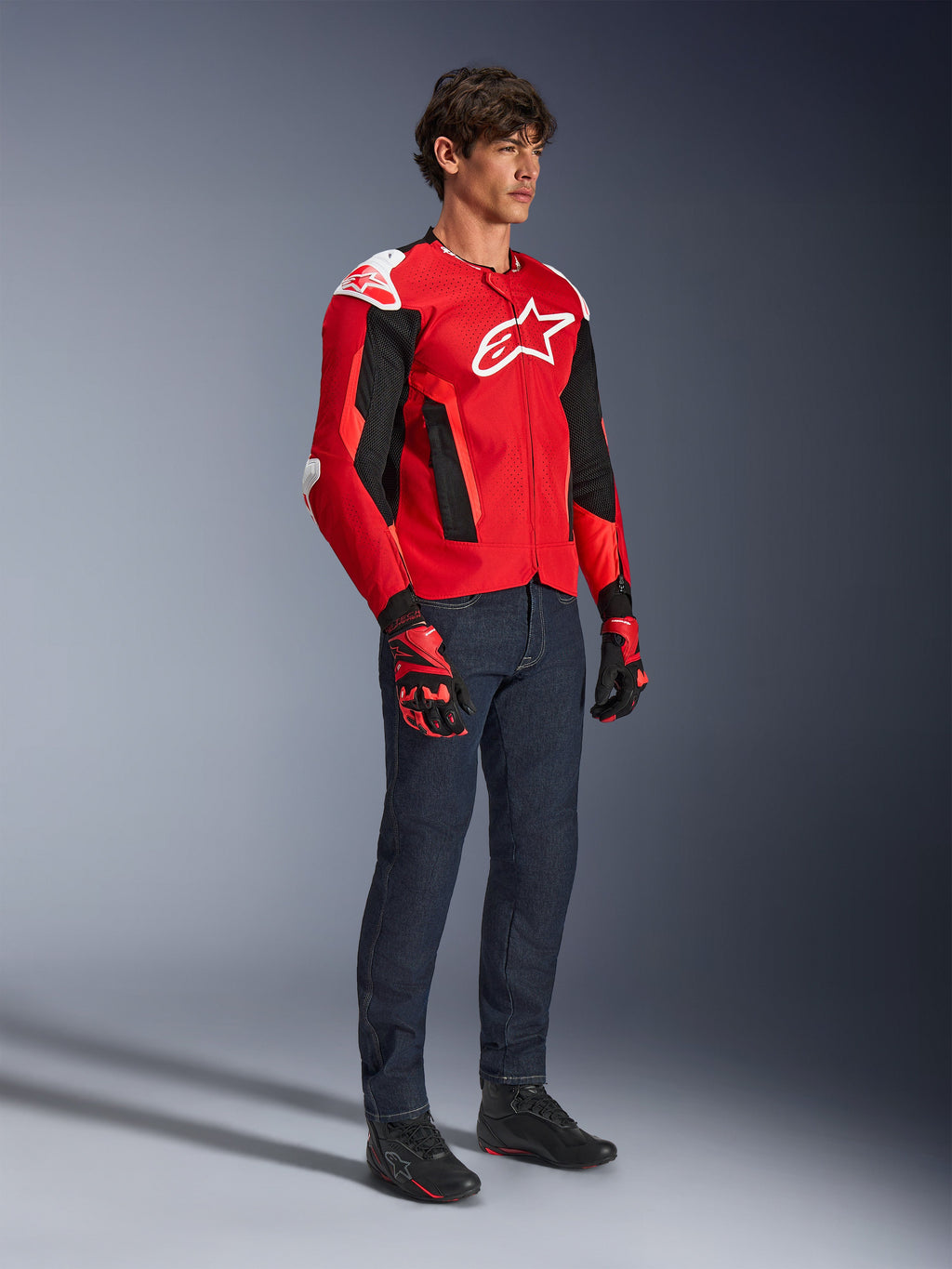 Alpinestars T-GP Air Jacket, sportieve motorjas, helderrood en zwart met witte accenten, gedragen door een model met een donkerblauwe jeans en rode racehandschoenen, voorzien van high-flow mesh-panelen en witte schoudersliders, aerodynamische sportpasvorm voor rijden in warm weer