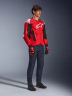 Alpinestars T-GP Air Jacket, sportieve motorjas, helderrood en zwart met witte accenten, gedragen door een model met een donkerblauwe jeans en rode racehandschoenen, voorzien van high-flow mesh-panelen en witte schoudersliders, aerodynamische sportpasvorm voor rijden in warm weer