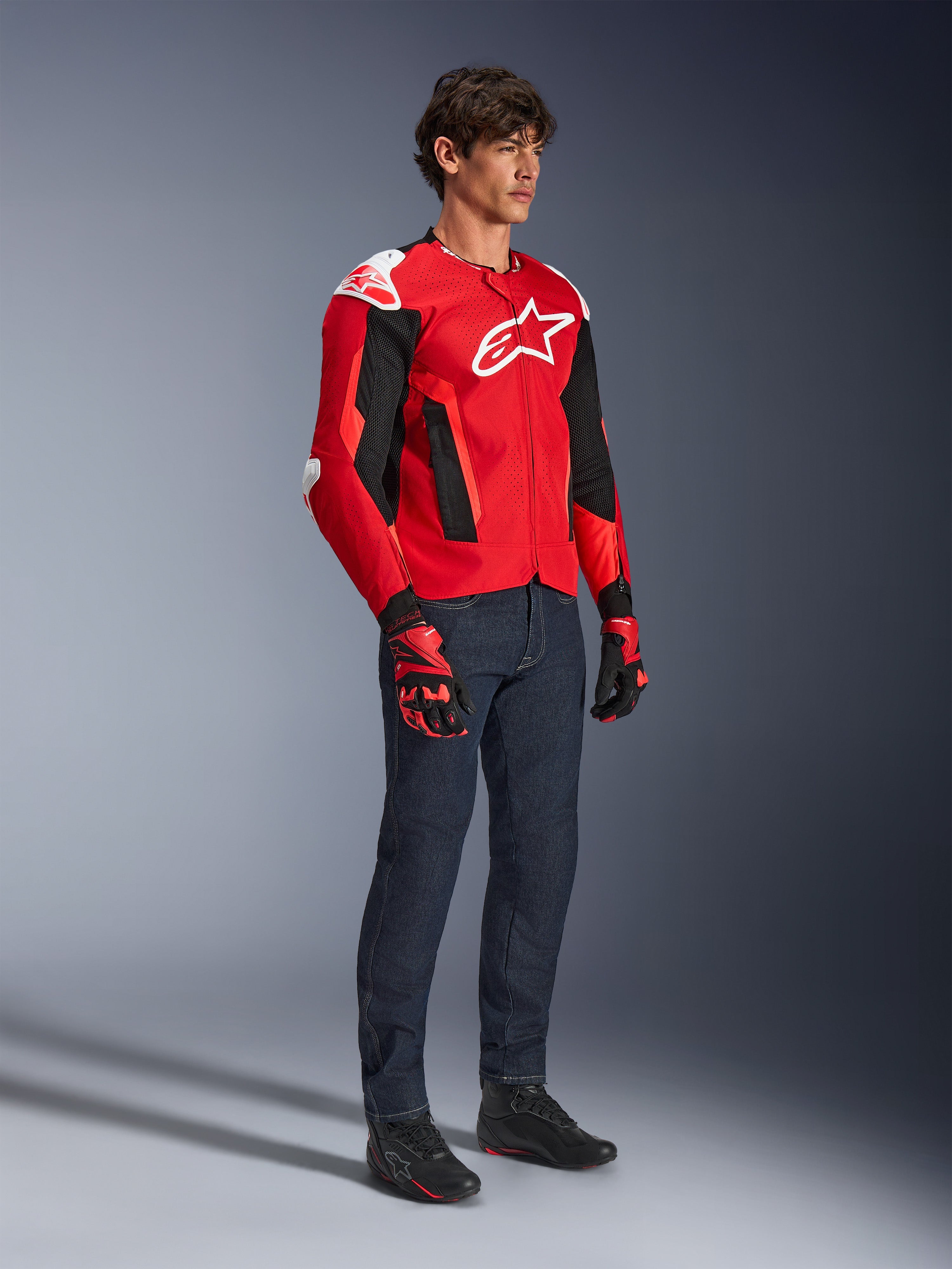 Alpinestars T-GP Air Jacket, sportieve motorjas, helderrood en zwart met witte accenten, gedragen door een model met een donkerblauwe jeans en rode racehandschoenen, voorzien van high-flow mesh-panelen en witte schoudersliders, aerodynamische sportpasvorm voor rijden in warm weer