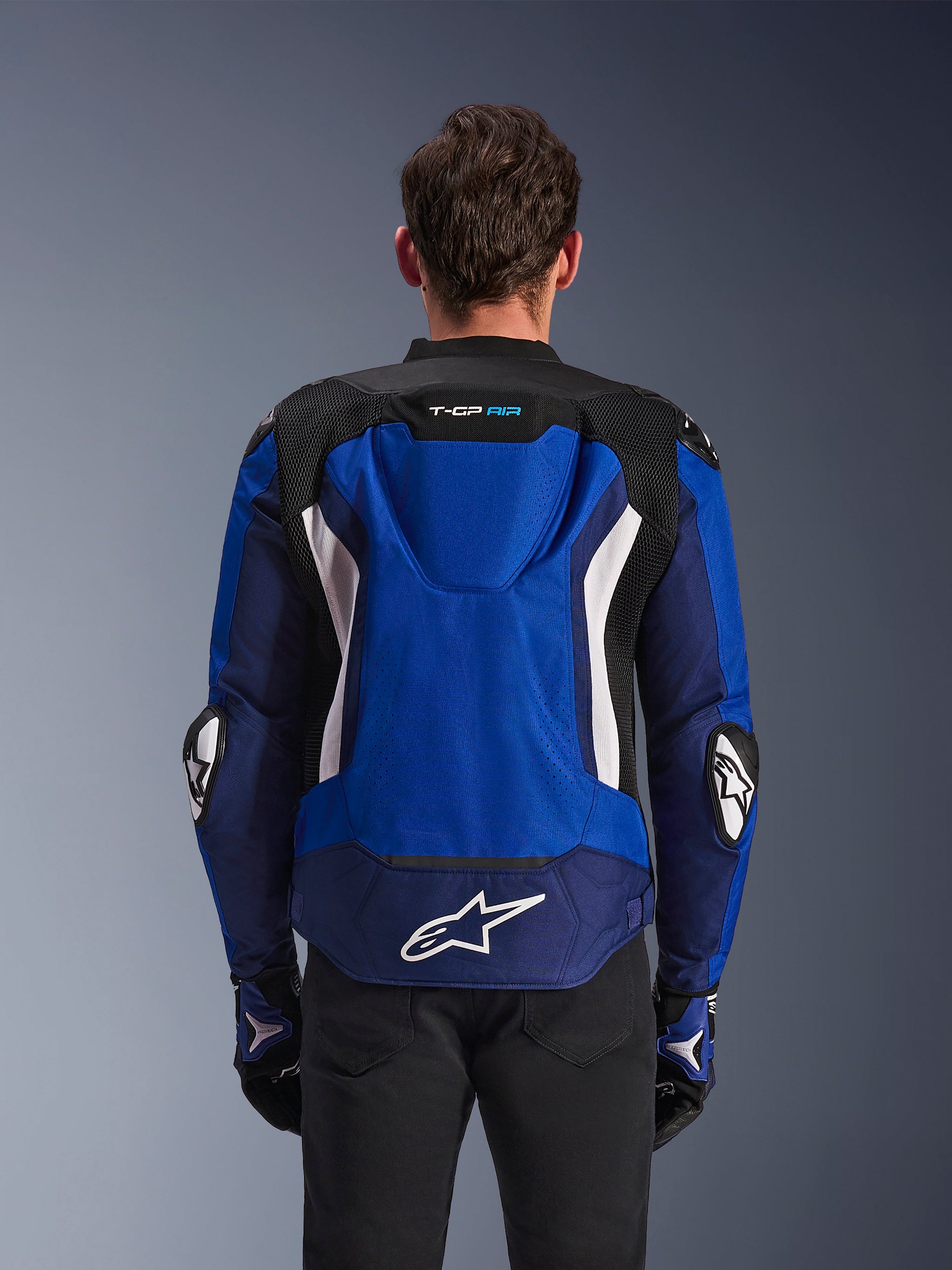 Alpinestars T-GP Air Jacket, sportieve textiele motorjas, helderblauw, navy en wit, achteraanzicht met de geïntegreerde aerodynamische bult en een groot wit logo op de onderrug, gedragen met bijpassende blauwe en zwarte motorhandschoenen