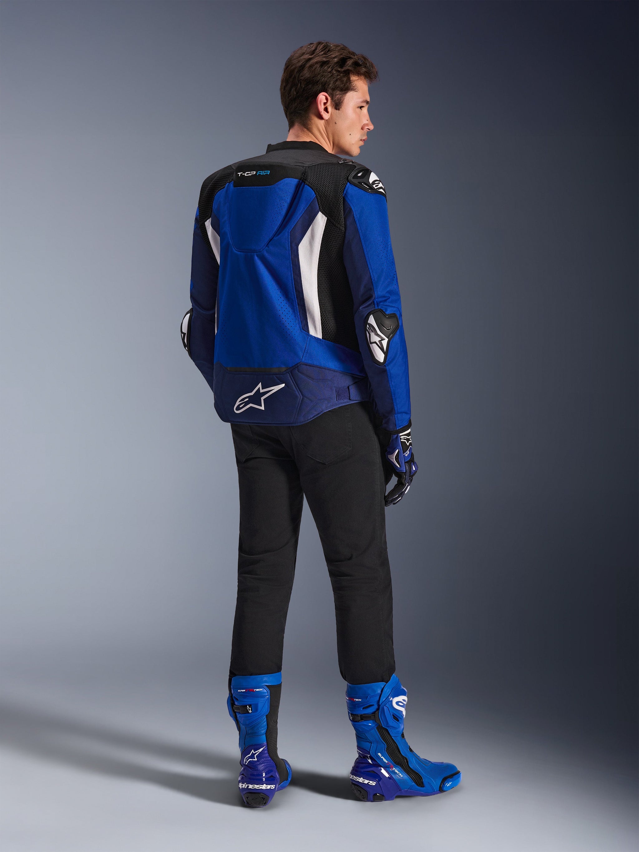 Alpinestars T-GP Air Jacket, sportieve textieljas, helderblauw, navy en wit, van achteren gezien op een model met een zwarte broek en bijpassende blauw-witte laarzen, voorzien van een wit Alpinestars-logo op de onderrug, mesh ventilatiepanelen en een aerodynamische bult