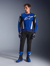 Mannelijk model in de Alpinestars T-GP Air Jas in Bright Blue Navy White, gecombineerd met een zwarte broek, bijpassende blauwe en marineblauwe racehandschoenen en blauwe en zwarte motorlaarzen, studiofoto van een complete sportieve motoruitrusting.