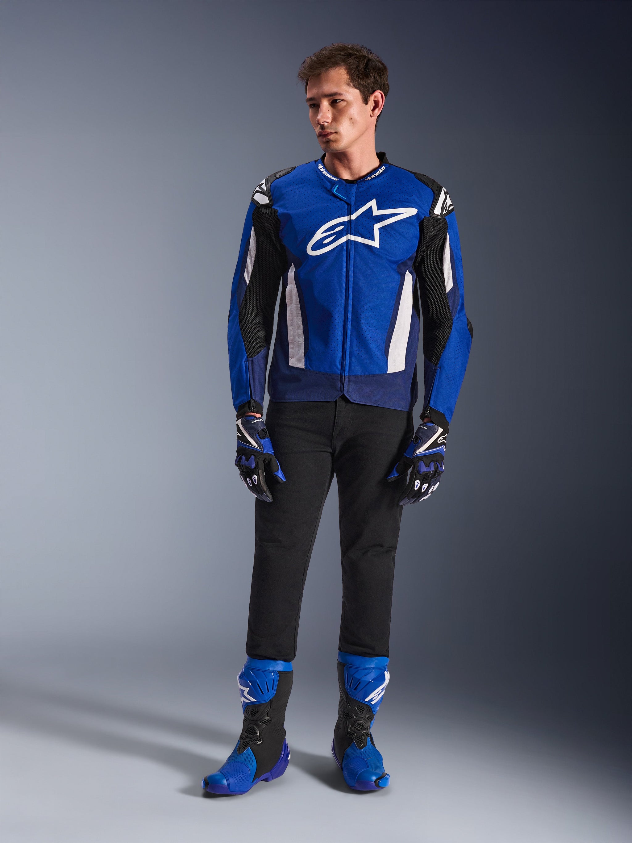 Mannelijk model in de Alpinestars T-GP Air Jas in Bright Blue Navy White, gecombineerd met een zwarte broek, bijpassende blauwe en marineblauwe racehandschoenen en blauwe en zwarte motorlaarzen, studiofoto van een complete sportieve motoruitrusting.