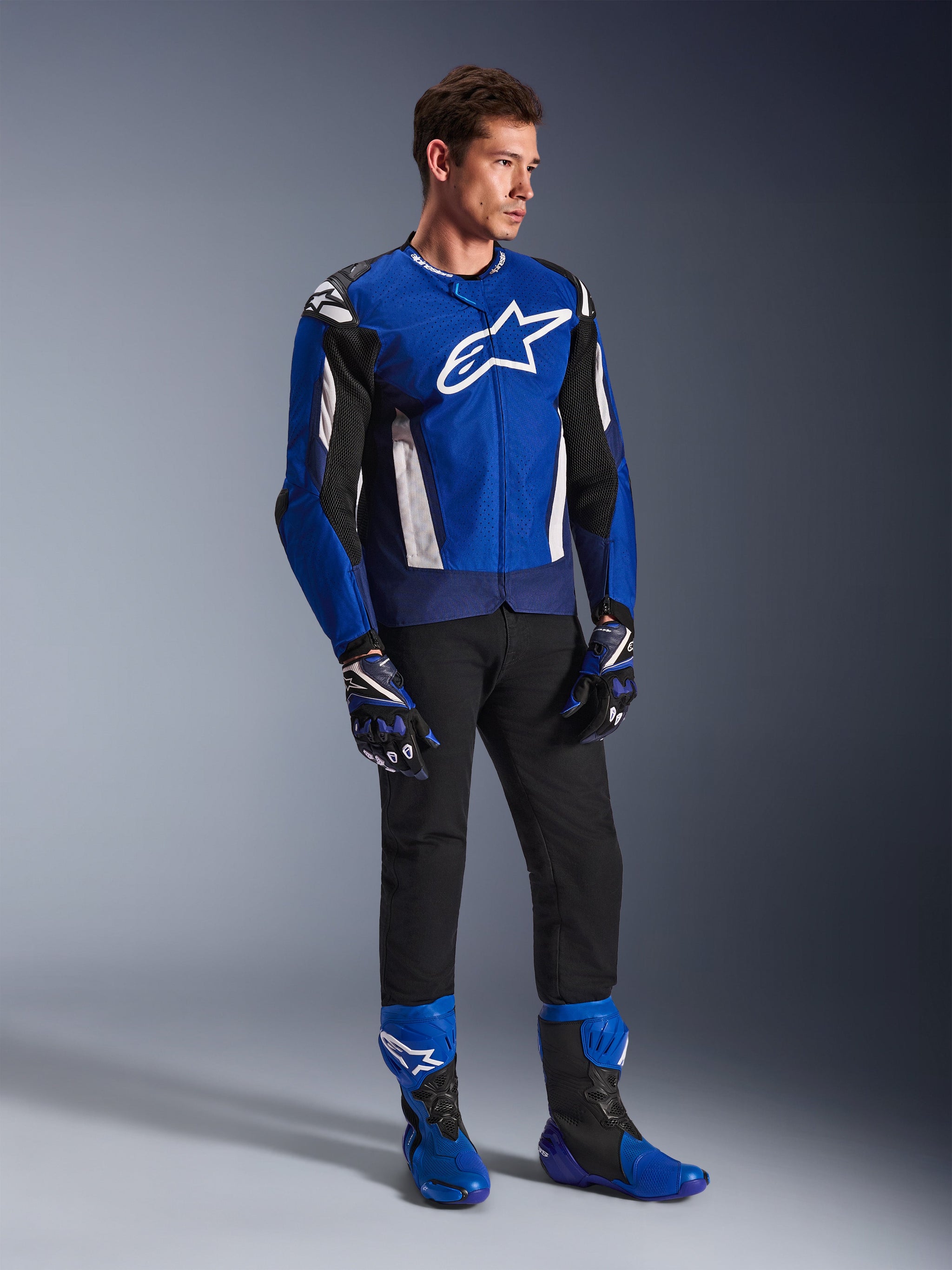 Alpinestars T-GP Air Jacket, sportieve textiele motorjas, helderblauw, marineblauw en wit, gedragen met bijpassende blauwe en zwarte handschoenen, zwarte broek en blauwe racelaarzen, voorzien van wit logo op de borst en technische mesh-panelen, studio-opname van het volledige lichaam