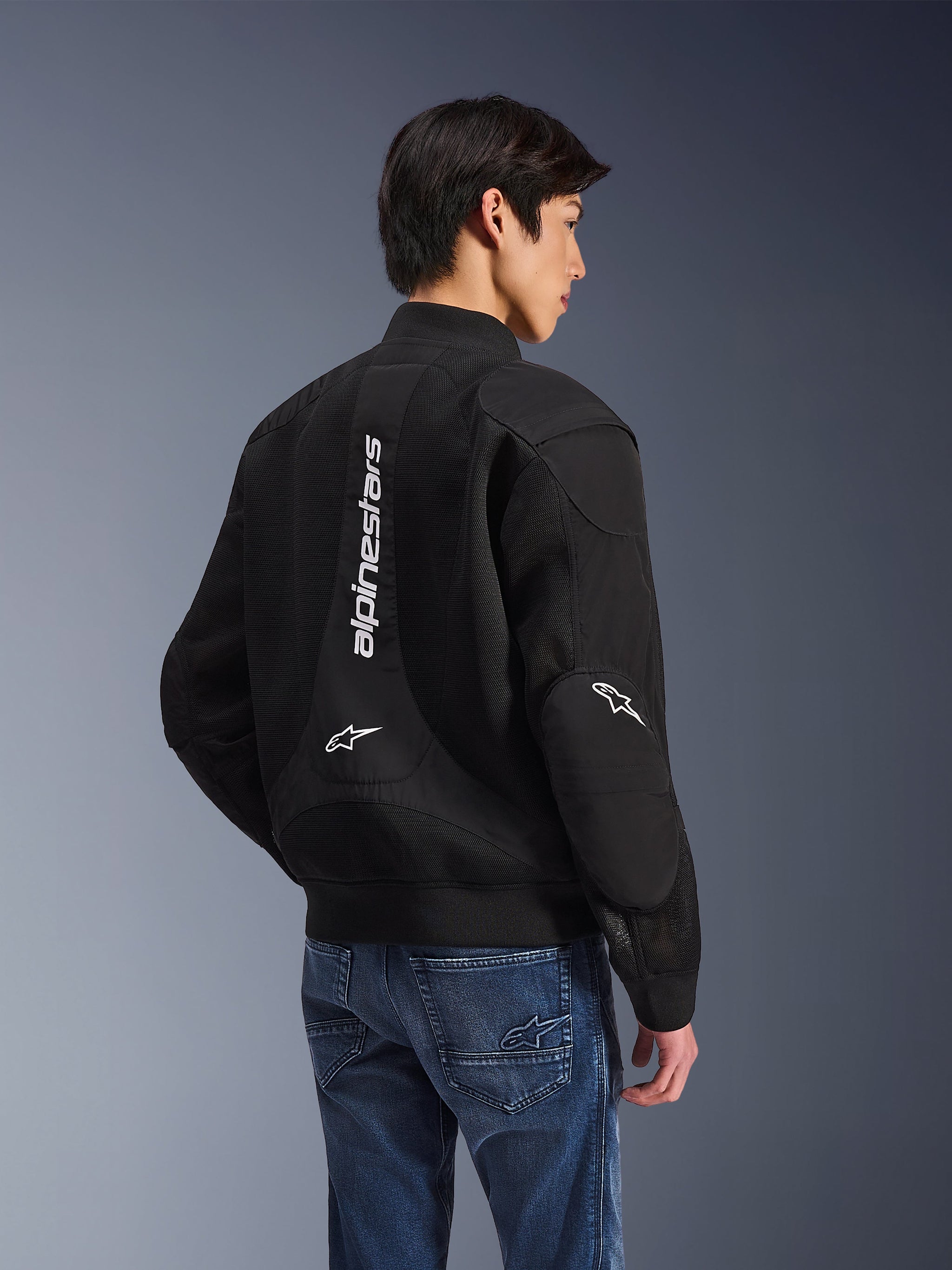 Alpinestars Flight Air Jacket, zwarte motorjas, achteraanzicht met groot verticaal wit logo en mesh ventilatiepanelen, bomber-stijl met geribbelde zoom, gedragen met blauwe jeans, urban contemporary motorkleding
