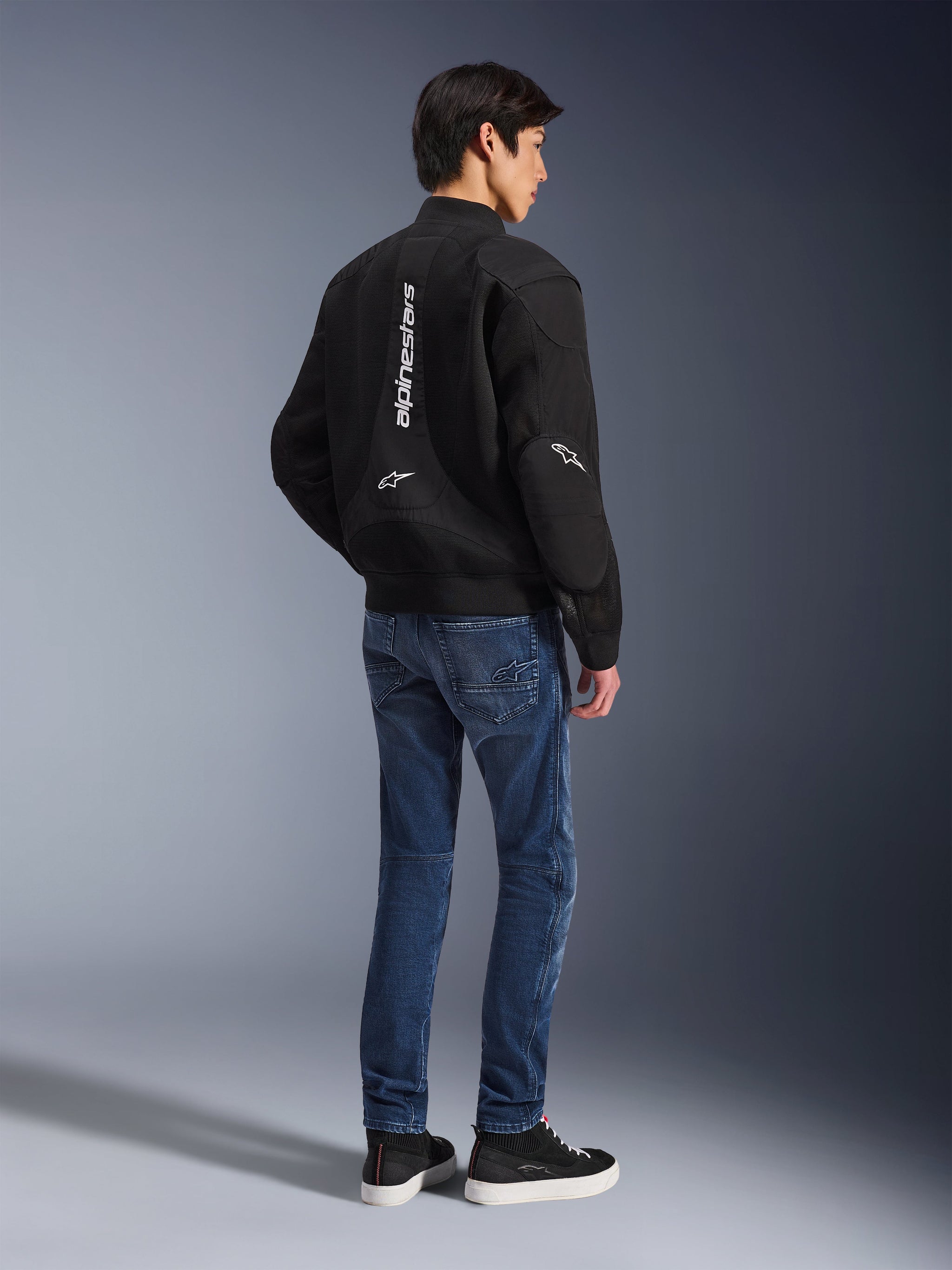 Model draagt Alpinestars Flight Air Jacket in het zwart, van achteren gezien, gecombineerd met een blauwe motorjeans en zwarte sneakers, met verticaal wit Alpinestars-logo en 3D-mesh ventilatiepanelen.