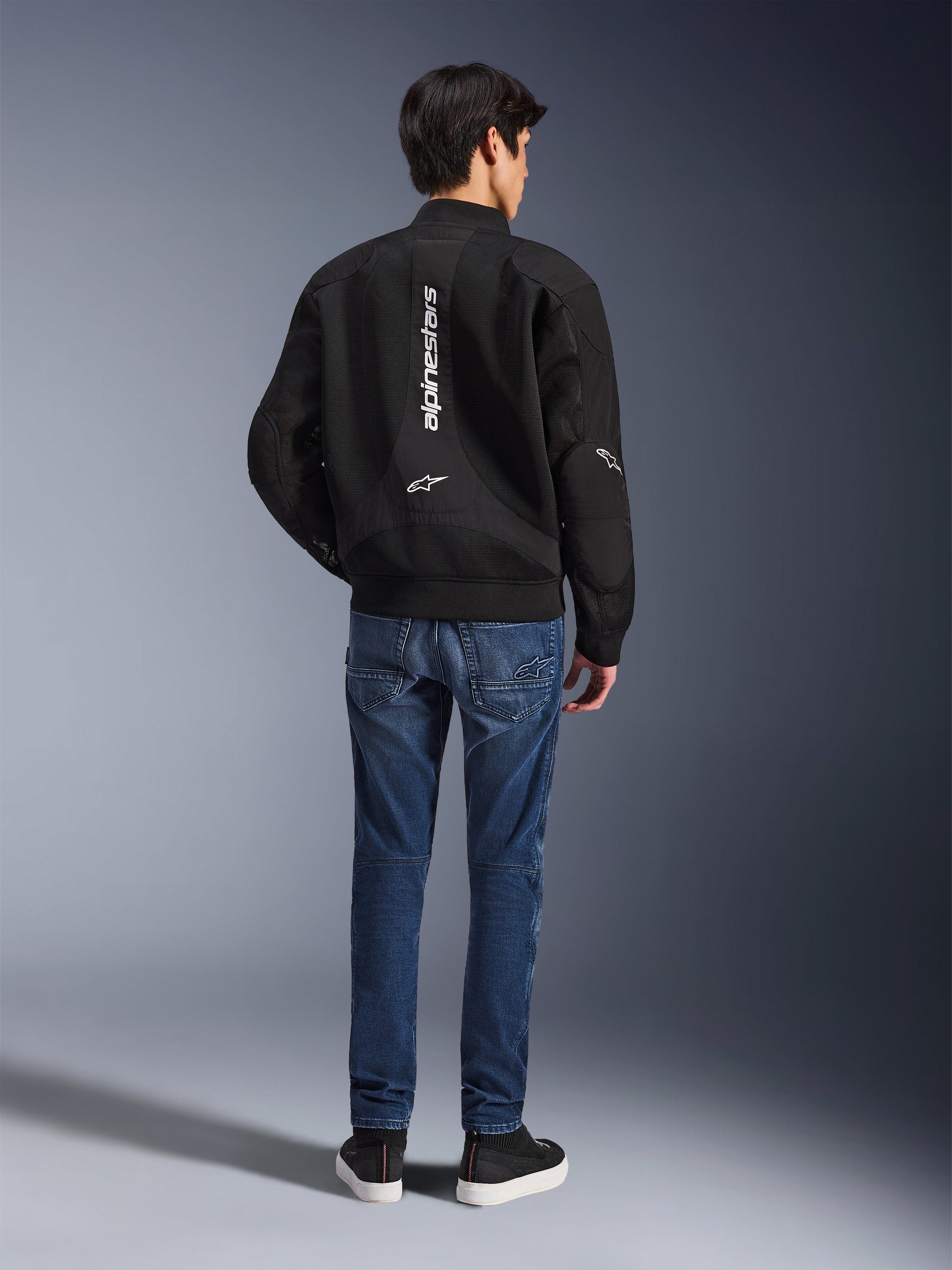 Model draagt de Alpinestars Flight Air Jacket van achteren gezien, zwarte motorjas in bomber-stijl met 3D mesh-panelen en een verticaal wit logo, gecombineerd met een blauwe denim motorjeans en zwarte sneakers, moderne urban motoruitrusting