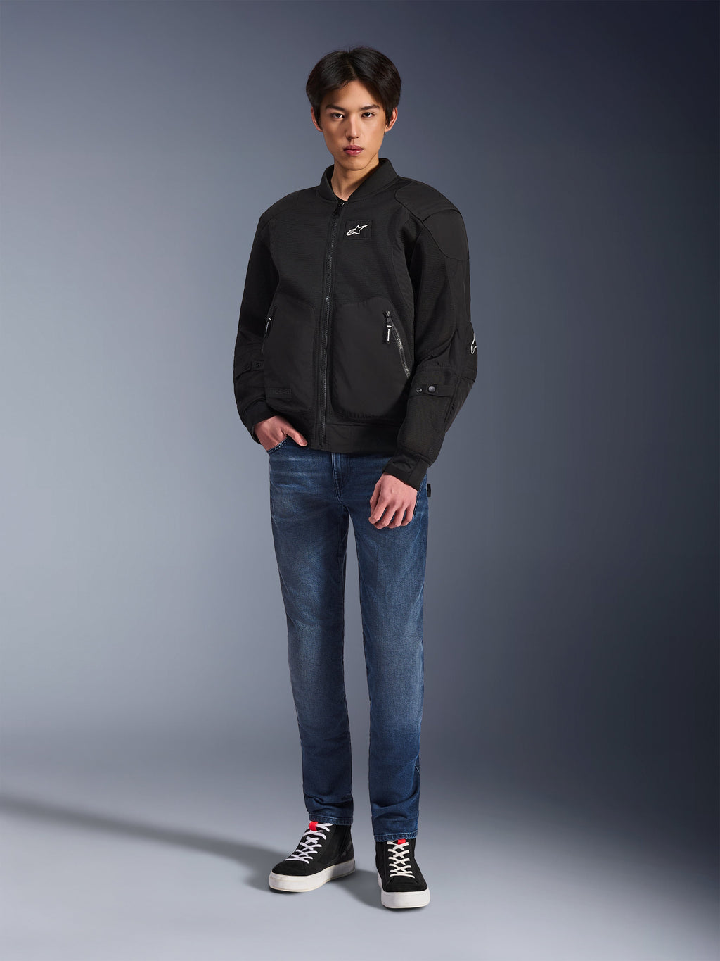 Model draagt de Alpinestars Flight Air Jacket in het zwart, een urban motorjas in bomberstijl met 3D mesh-panelen en een ritssluiting aan de voorzijde, gecombineerd met een blauwe jeans en zwarte sneakers, eigentijdse op streetstyle geïnspireerde motorkleding.