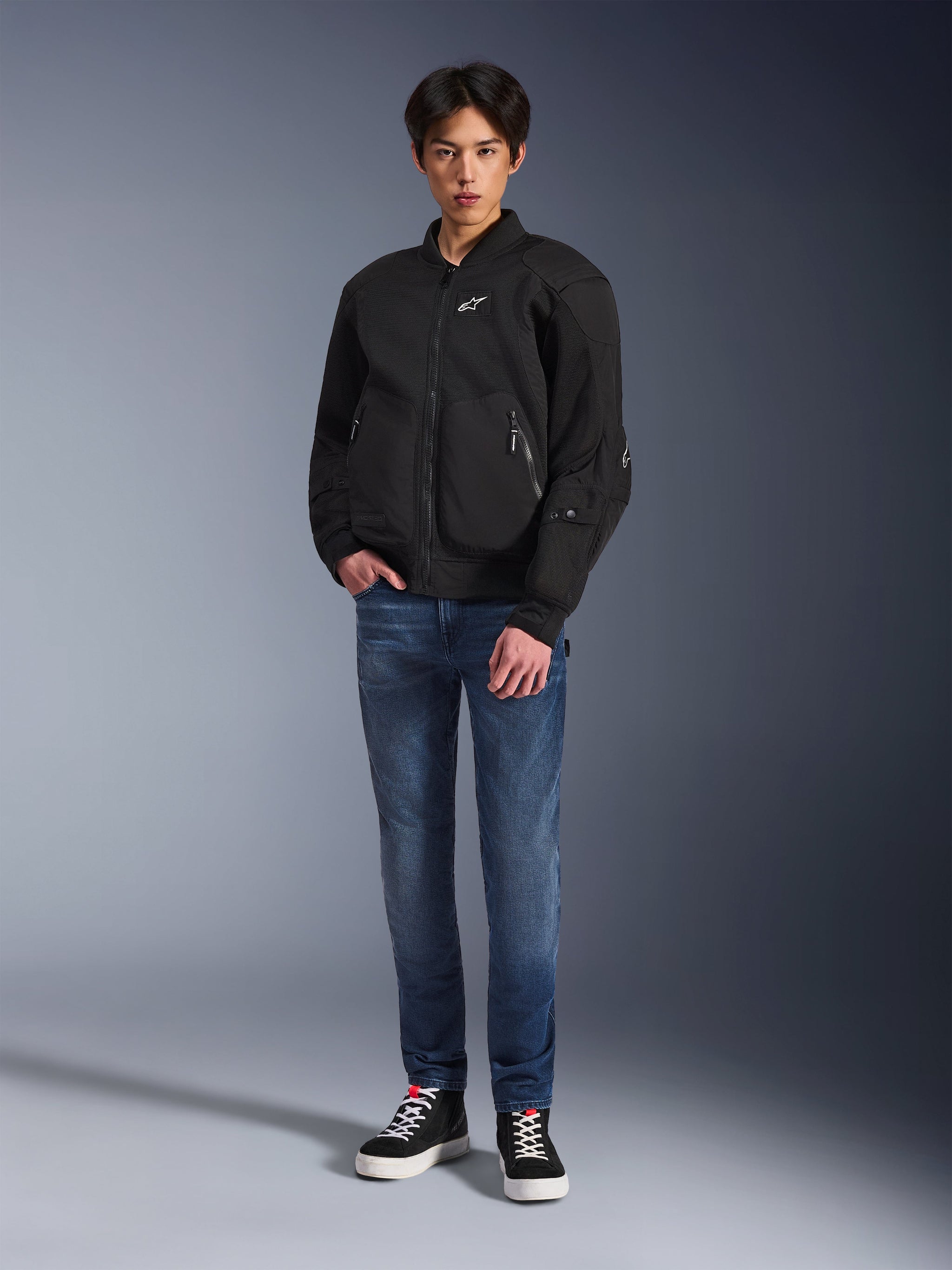 Model draagt de Alpinestars Flight Air Jacket in het zwart, een urban motorjas in bomberstijl met 3D mesh-panelen en een ritssluiting aan de voorzijde, gecombineerd met een blauwe jeans en zwarte sneakers, eigentijdse op streetstyle geïnspireerde motorkleding.