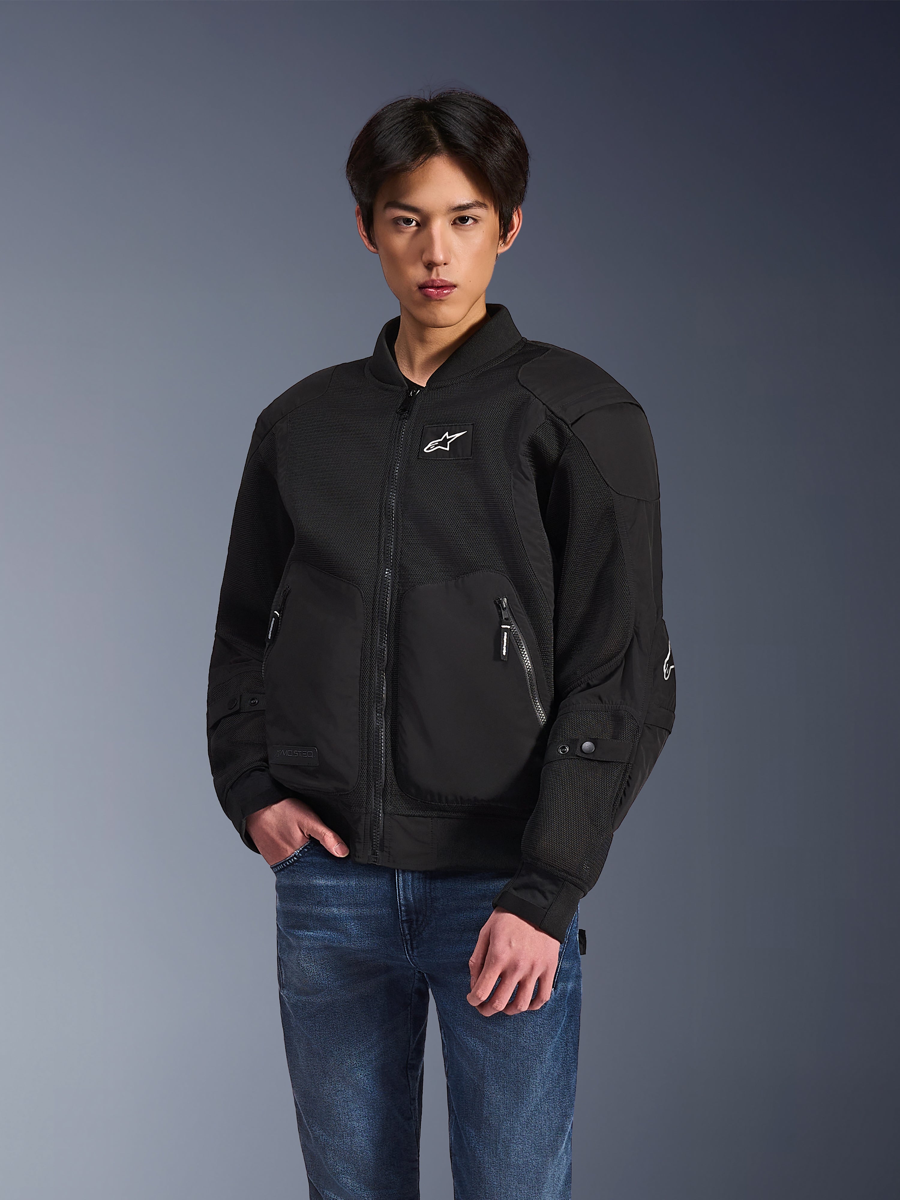 Alpinestars Flight Air Jacket, zwarte urban motorjas, gedragen door een man met blauwe jeans, met een bomberstijl-esthetiek met mesh-panelen, ritssluiting aan de voorzijde en wit logo op de borst, ontworpen voor stadsritten in warm weer