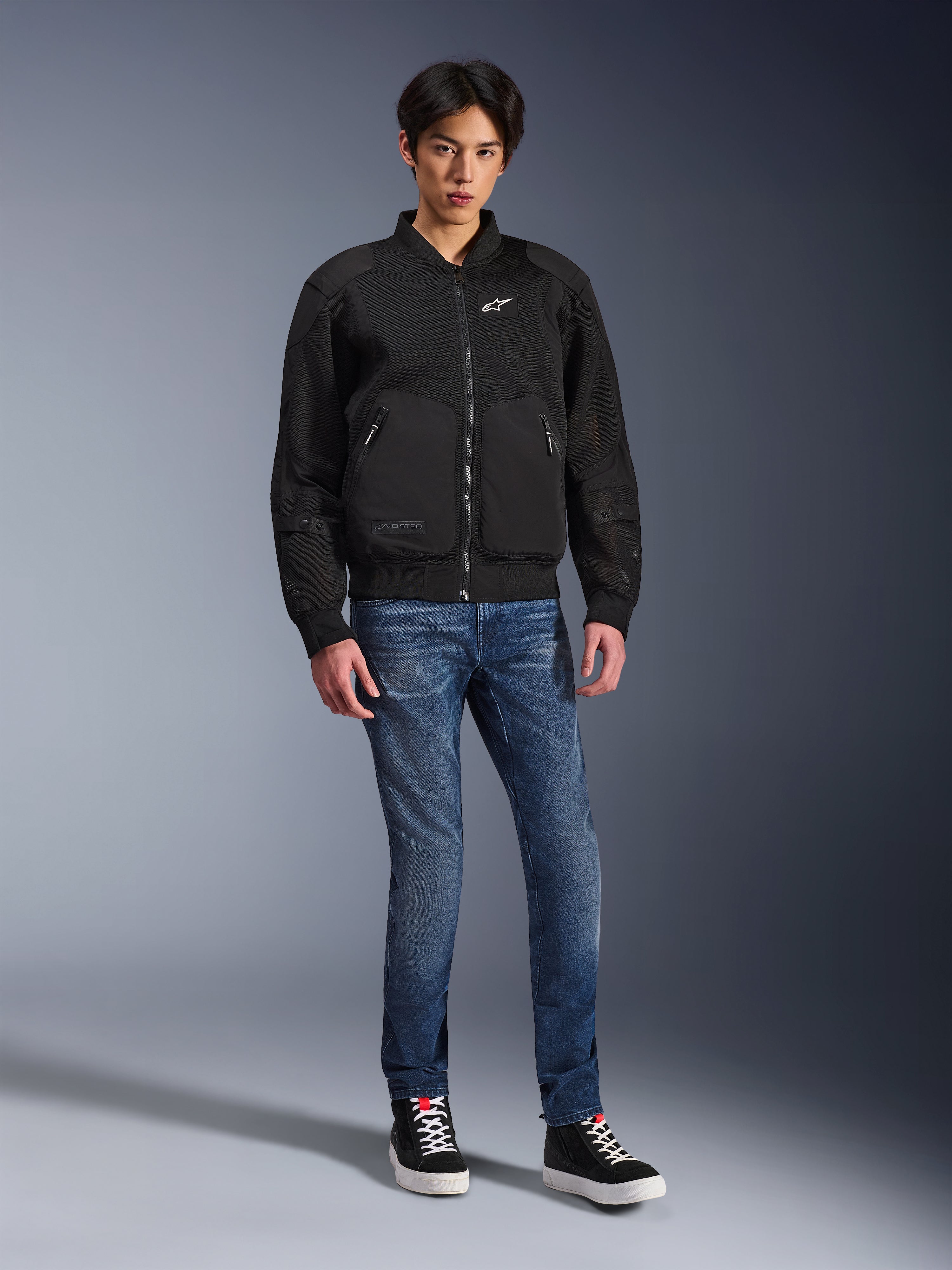 Alpinestars Flight Air Jacket, zwart, stedelijk geïnspireerde motorjas in bomberstijl met 3D mesh-panelen en wit logo op de borst, gedragen door een model met blauwe jeans en zwarte sneakers, eigentijdse motorkleding
