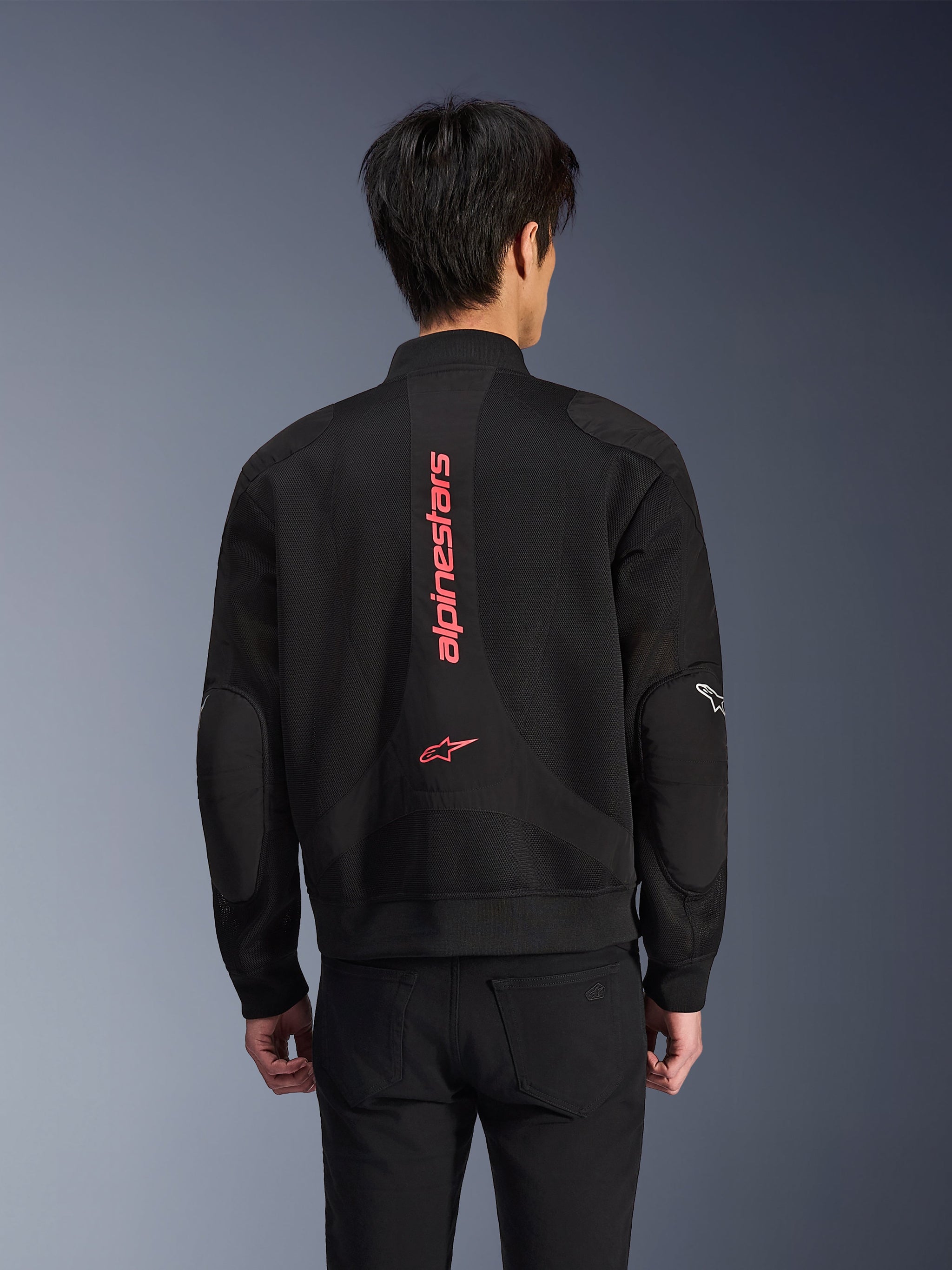 Achteraanzicht van een motorrijder die de Alpinestars Flight Air Jacket draagt in Black Bright Red, urban motorjas in bomberstijl met 3D-mesh-constructie, voorzien van groot verticaal rood Alpinestars-logo en sterlogo op de rug, technische motorkleding geïnspireerd op streetwear