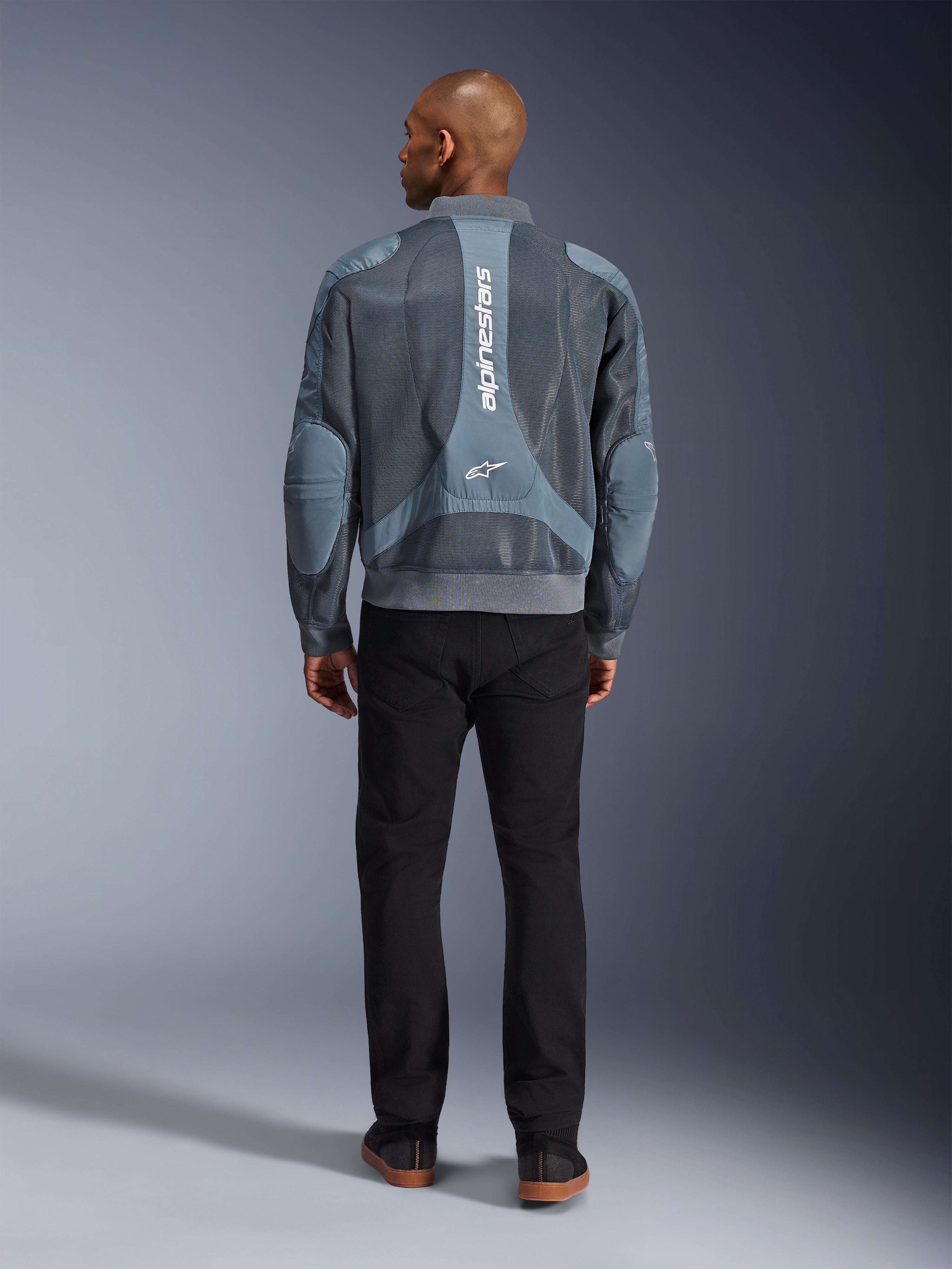 Alpinestars Flight Air Jacket in Storm Gray, achteraanzicht van een mannelijk model dat een urban-stijl motorjas draagt met 3D-meshpanelen en verticale Alpinestars-branding, gecombineerd met een zwarte broek voor een moderne forenzenlook