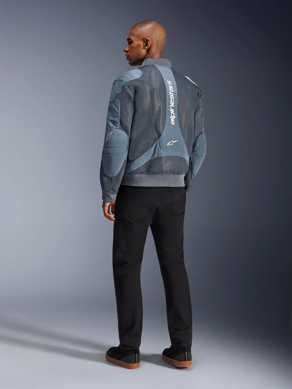 Alpinestars Flight Air Jacket, Storm Gray motorjas, achteraanzicht, voorzien van 3D mesh ventilatiepanelen en verticale witte Alpinestars-branding, gedragen met zwarte broek en zwarte sneakers, urban commuter-stijl