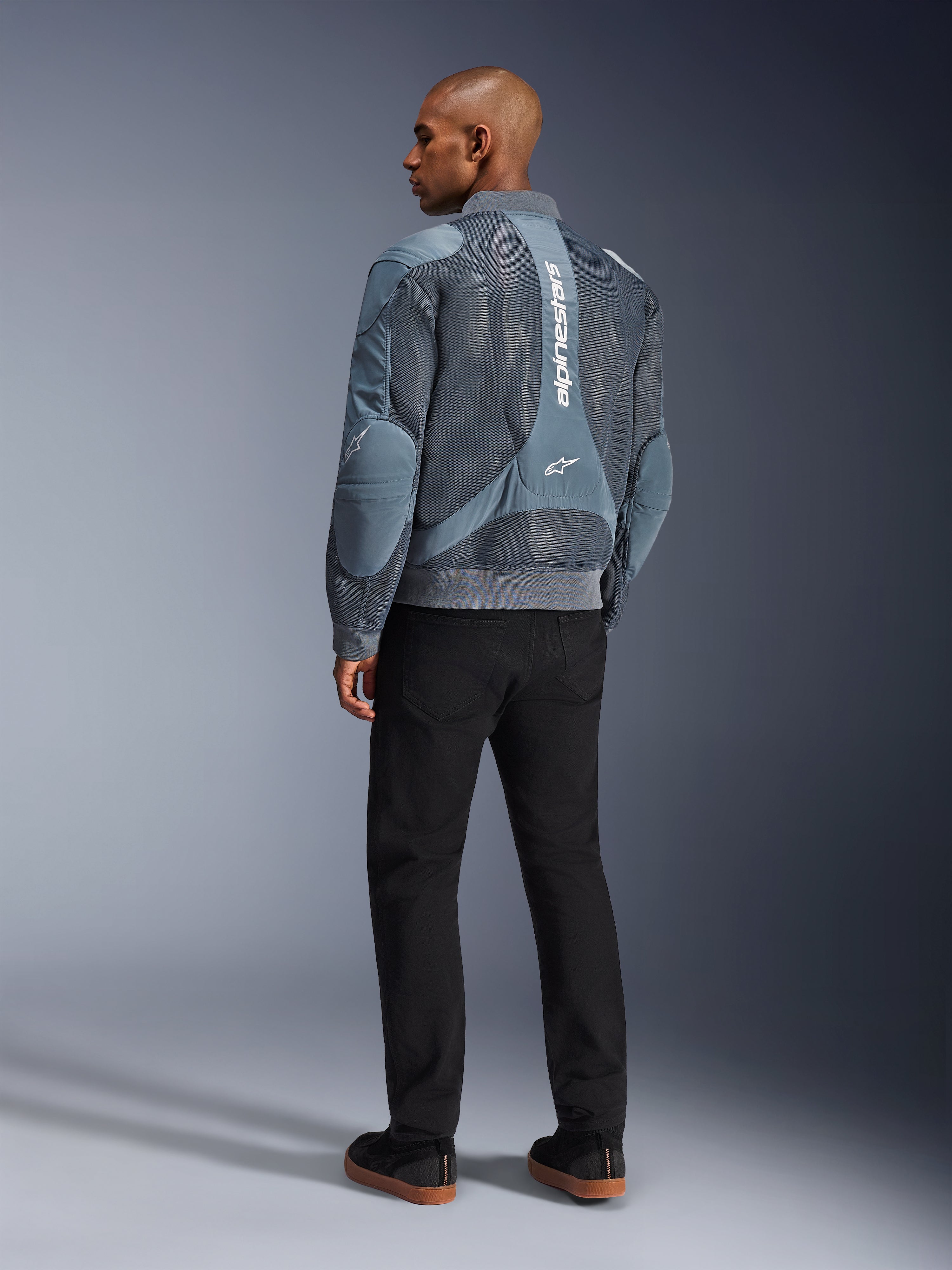 Alpinestars Flight Air Jacket, Storm Gray motorjas, achteraanzicht, voorzien van 3D mesh ventilatiepanelen en verticale witte Alpinestars-branding, gedragen met zwarte broek en zwarte sneakers, urban commuter-stijl