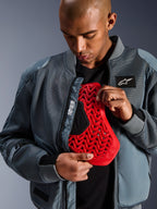 Alpinestars Flight Air Jacket in stormgrijs, urban motor bomberjack met 3D mesh-panelen voor optimale luchtstroom, opengeritst om een rode Nucleon rugprotector-insert te tonen, voorzien van 'Alpinestars Protects' branding en een moderne streetwear-esthetiek