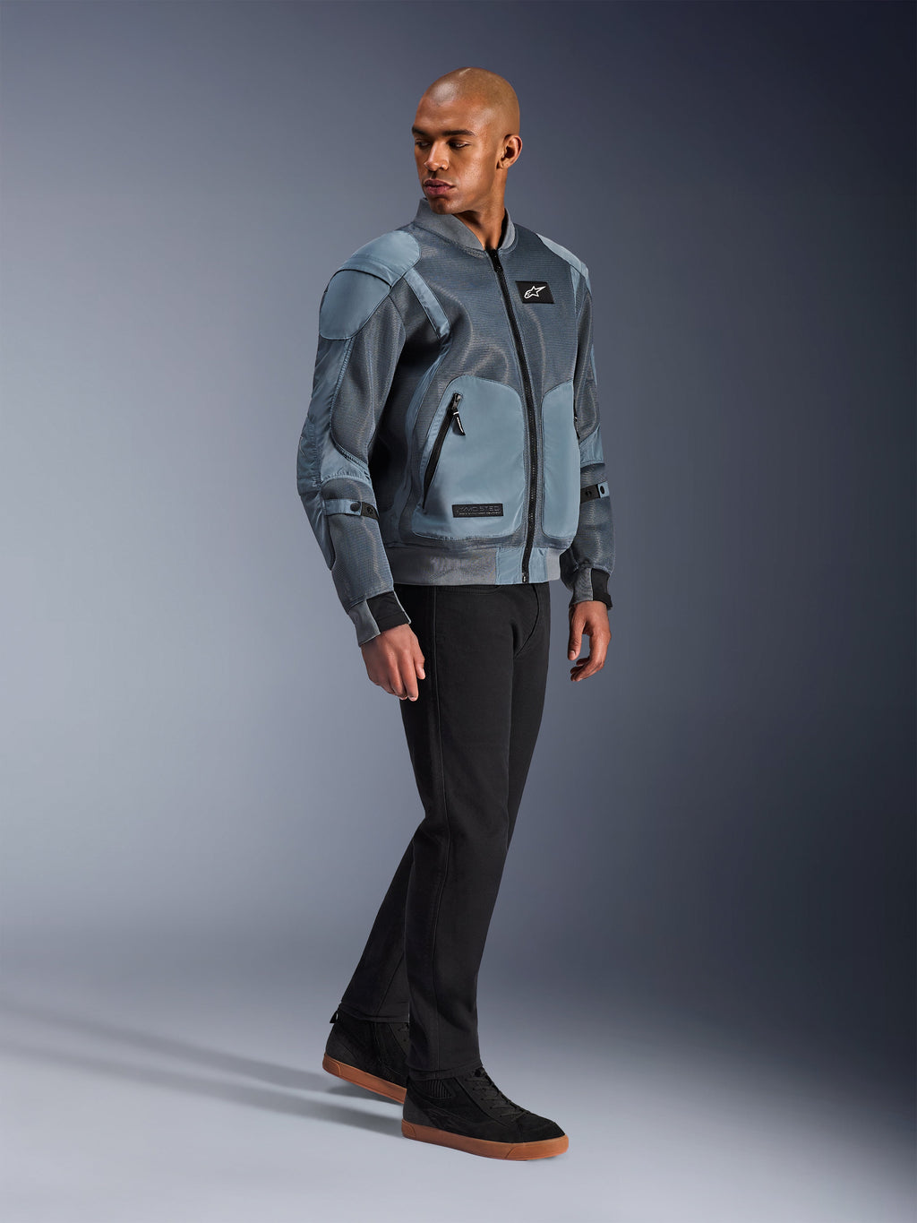 Alpinestars Flight Air Jacket, Storm Gray, op urban bomber geïnspireerde motorjas, gedragen door een mannelijk model met zwarte broek en sneakers, voorzien van uitgebreide 3D-mesh ventilatiepanelen en externe protectorzakken, moderne streetwear beschermende kleding voor rijden bij warm weer