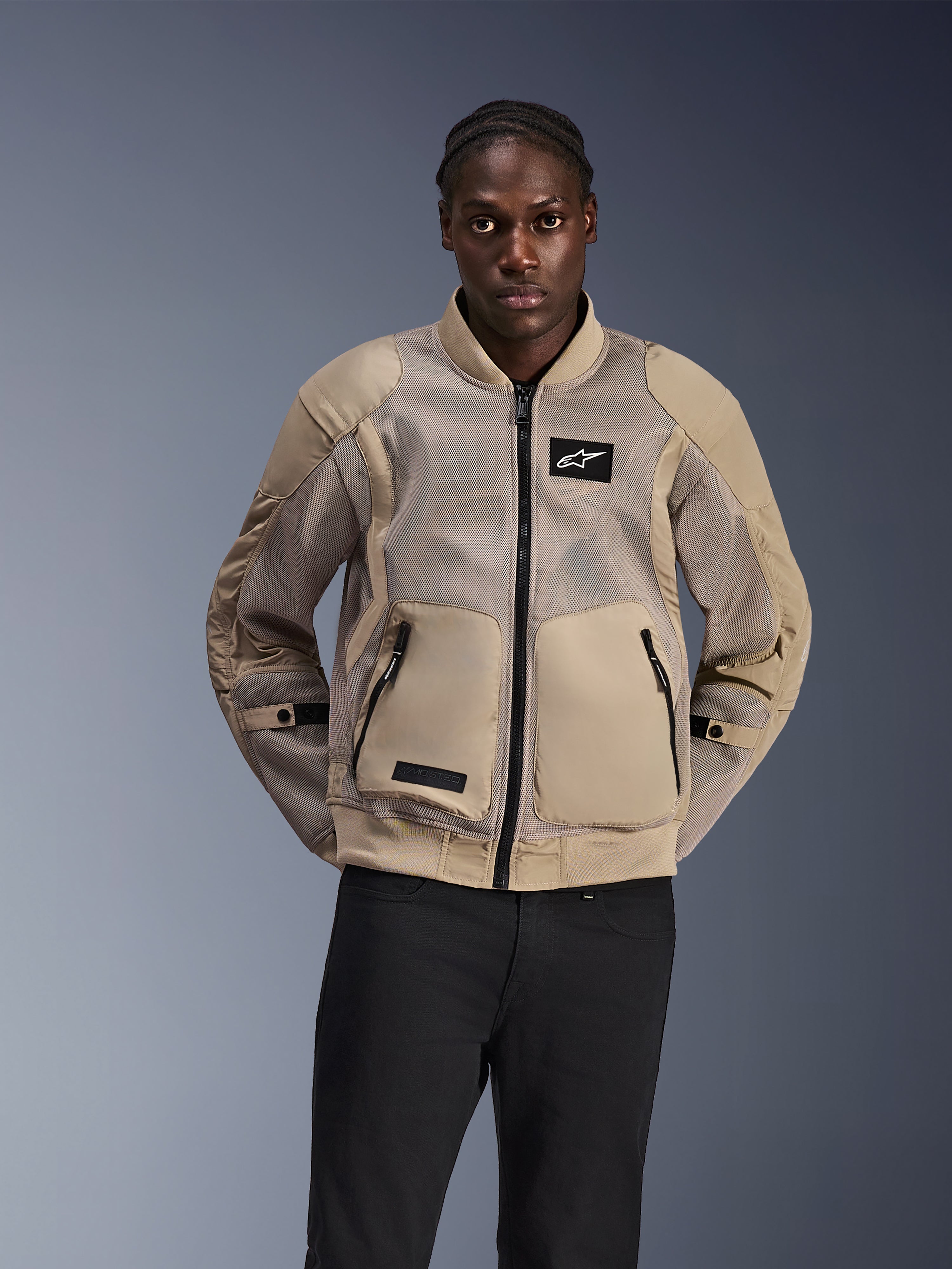Alpinestars Flight Air Jacket, stedelijke motorjas in bomberstijl, lichtbruin, voorzien van uitgebreide 3D-meshpanelen voor ventilatie en grote voorzakken, gedragen door een man met een zwarte broek, modern op motorsport geïnspireerd ontwerp voor ritten bij warm weer.