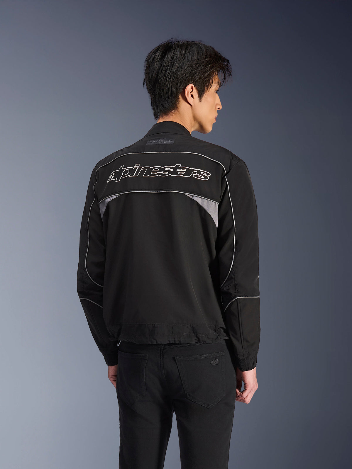 Alpinestars Unite Jas, urban motorjas, zwart met donkergrijze accenten, achteraanzicht met wit Alpinestars-woordmerk over de schouders, reflecterende biezen en panelen, minimalistisch softshell streetwear ontwerp