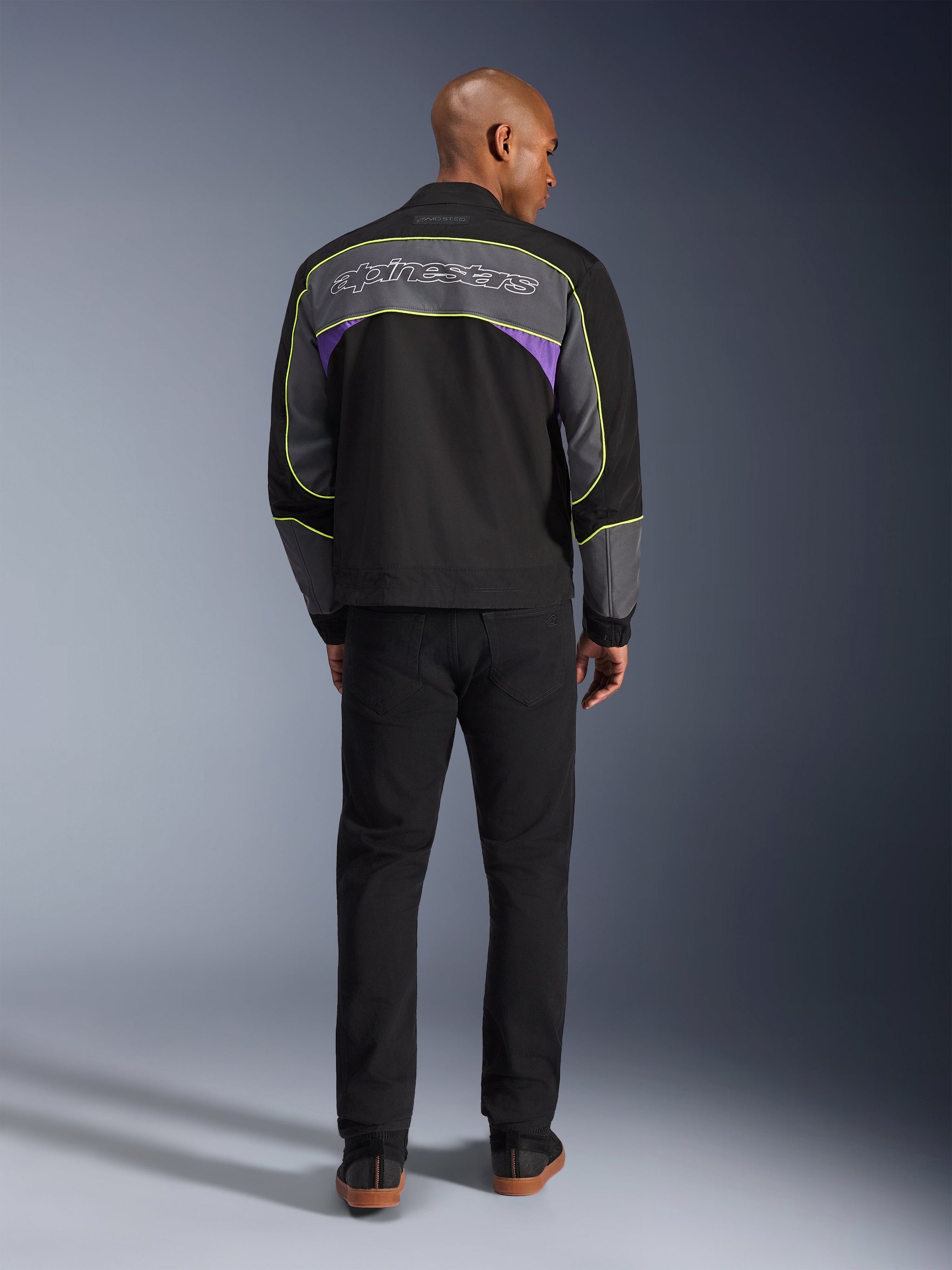 Alpinestars Unite Jacket, urban motorjas in Black Asphalt Violet, achteraanzicht met grijze rugpanelen, paarse accenten en neongele piping, Alpinestars-scriptlogo over de schouders, gedragen met een zwarte broek