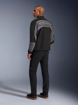Alpinestars Unite Jacket, urban motorjas, Black Asphalt Violet, driekwart achteraanzicht op model, zwarte softshell met grijze panelen en violette accenten, neongele reflecterende biezen, Alpinestars-logo op de bovenrug, gecombineerd met een zwarte broek en zwarte vrijetijdsschoenen