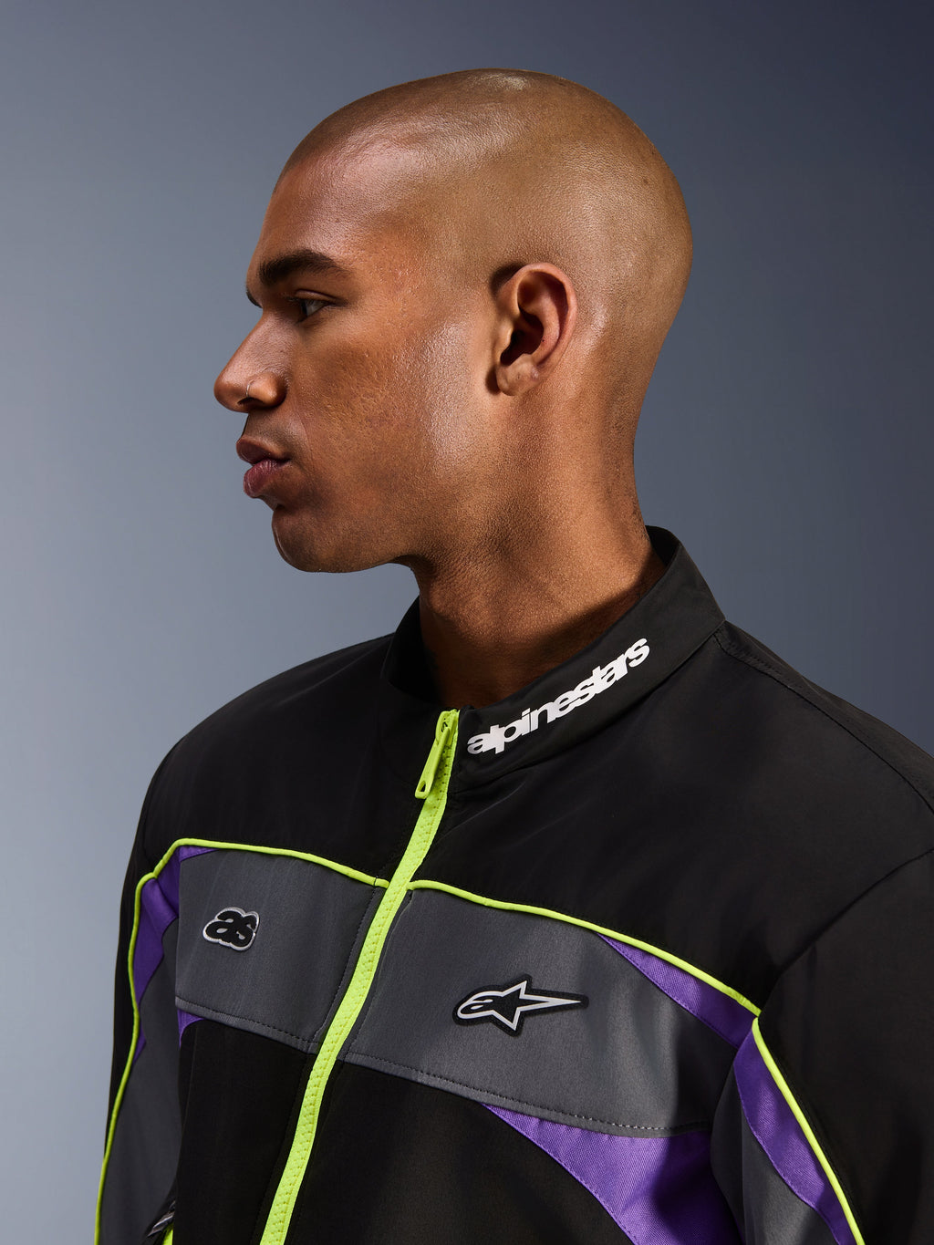 Alpinestars Unite Jacket, Black Asphalt Violet, profielaanzicht van mannelijk model met focus op de hoge kraag met witte Alpinestars-branding, neongele rits, grijze borstpanelen met logo's, paarse accenten, minimalistische urban motor-streetwear styling