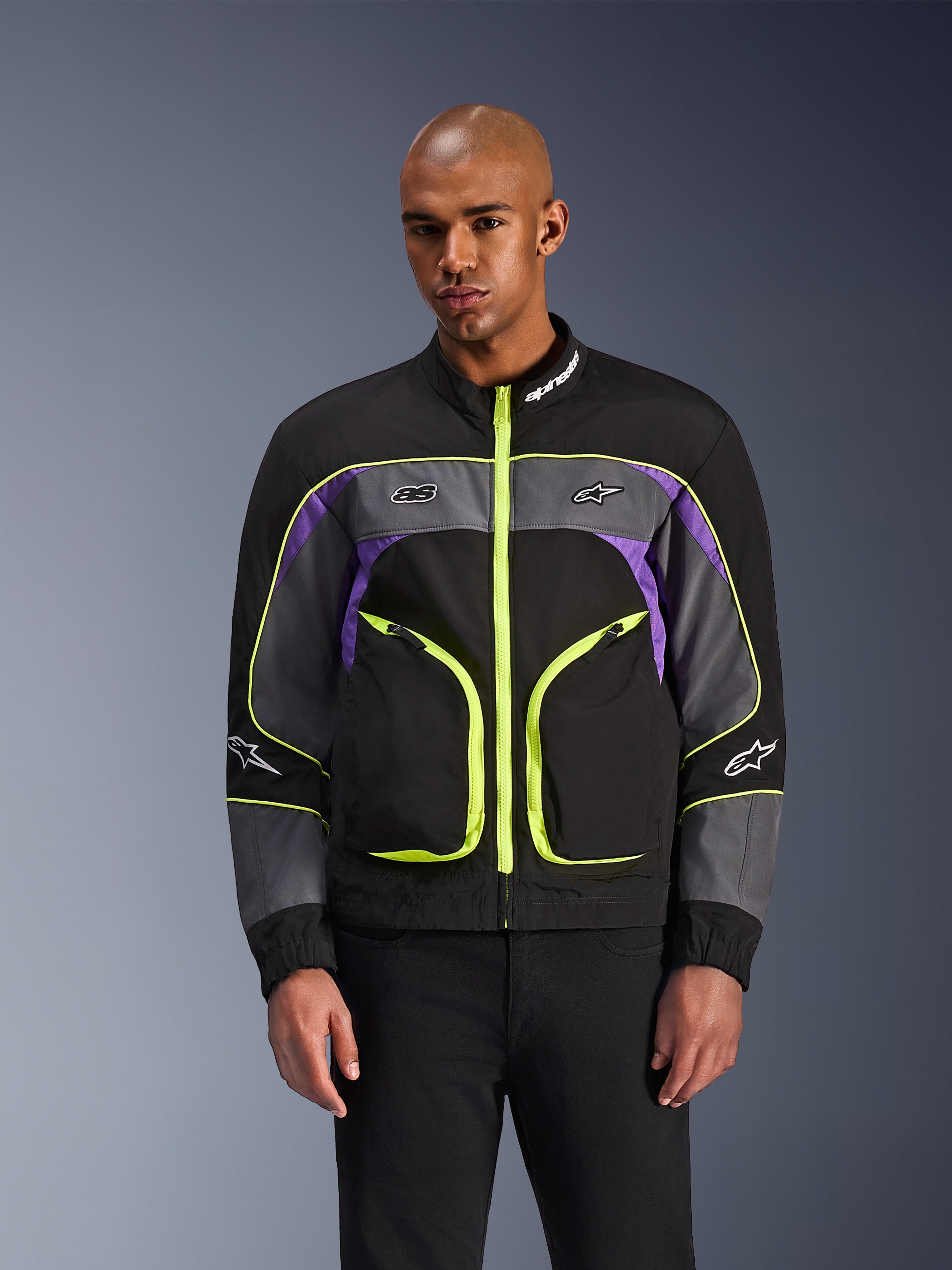 Alpinestars Unite Jacket, urban motorjas, Black Asphalt en Violet met neongele accenten, voorzien van grote voorzakken met neon ritsen, hoge kraag met witte merknaam, gedragen door een model, eigentijdse op streetwear geïnspireerde beschermende kleding
