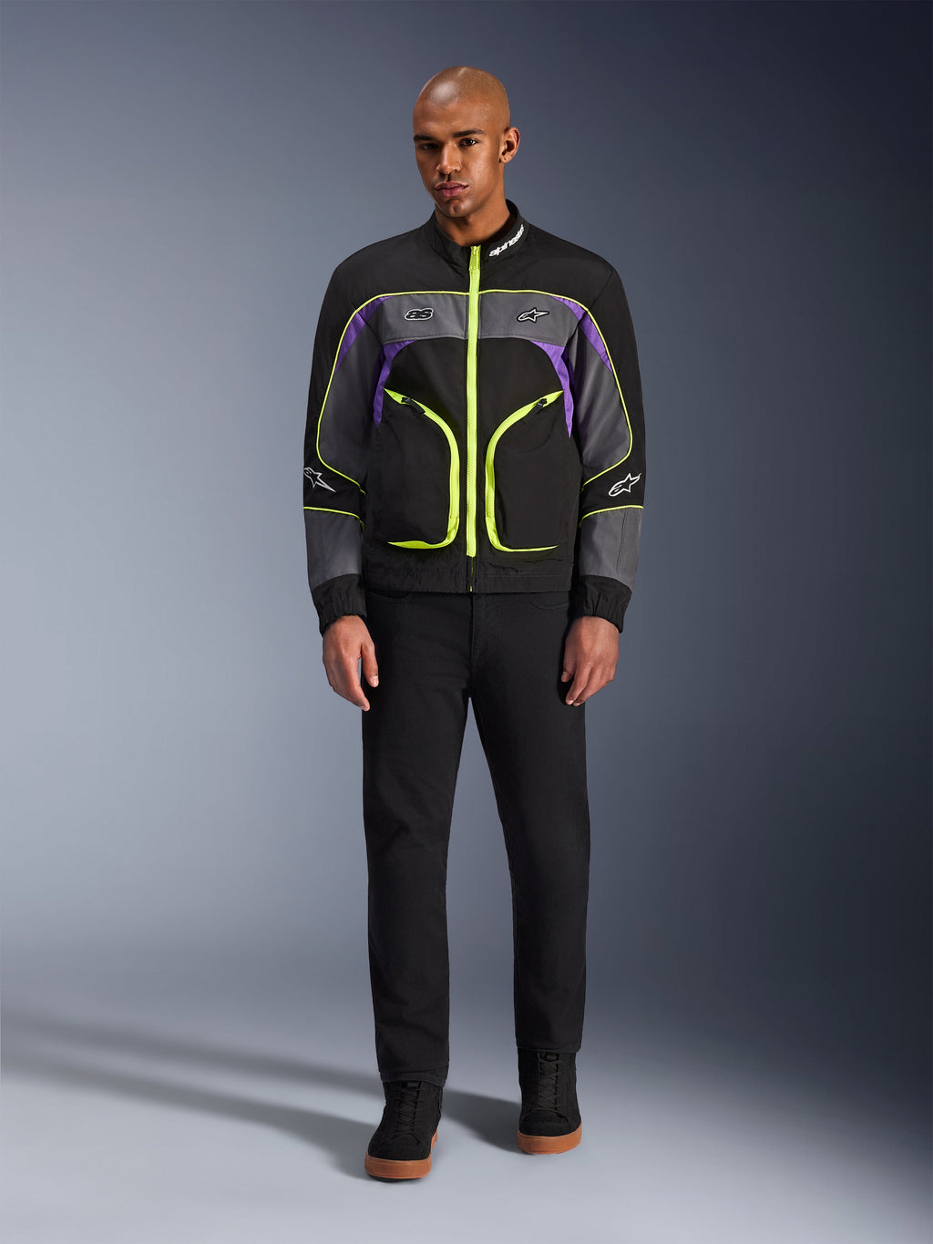 Mannelijk model draagt de Alpinestars Unite Jacket in Black Asphalt Violet, voorzien van een zwarte basis met grijze en paarse panelen en felle neongele ritsen en details, gecombineerd met een zwarte broek en schoenen, minimalistische urban motorjas.