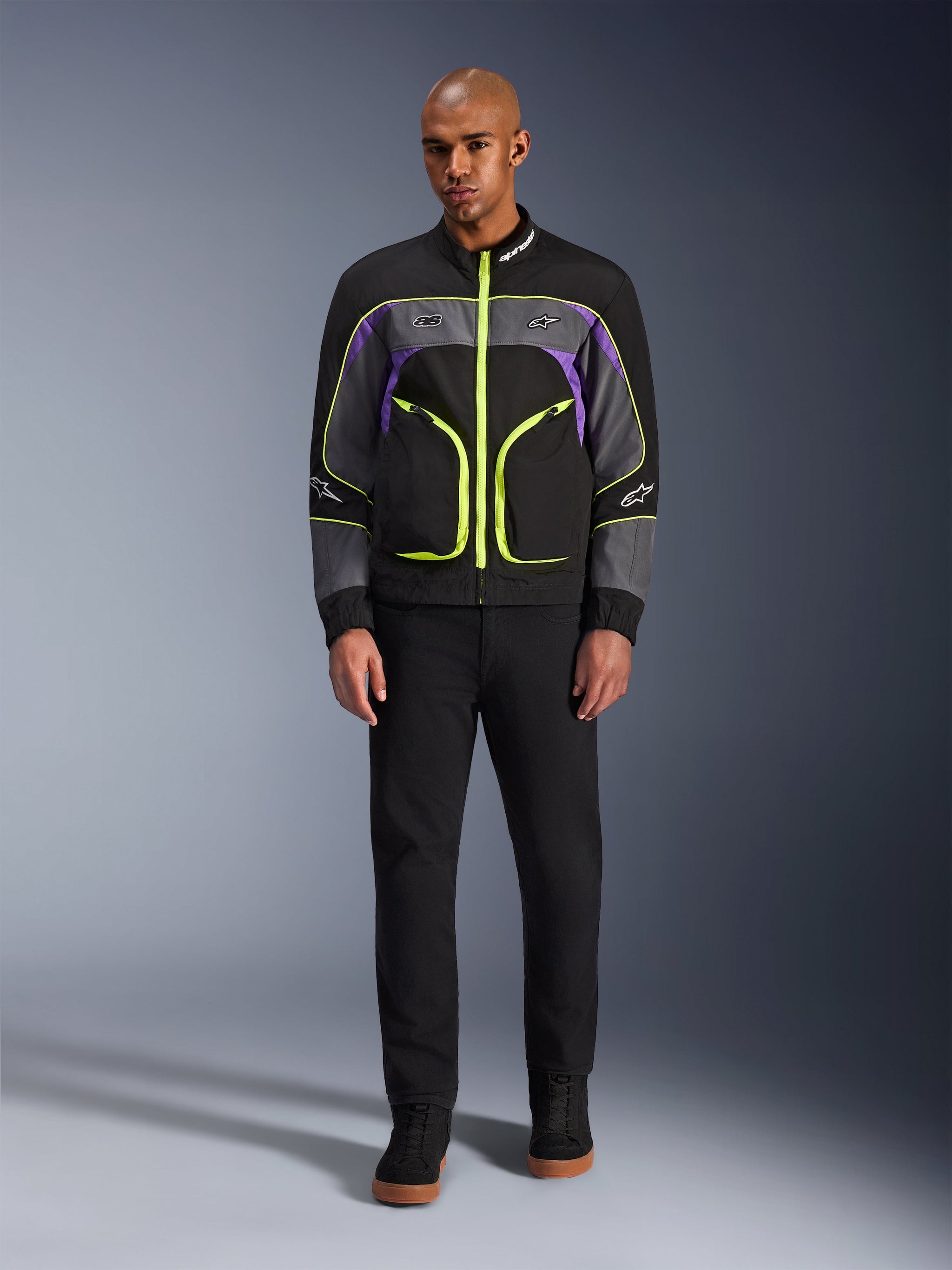 Mannelijk model draagt de Alpinestars Unite Jacket in Black Asphalt Violet, voorzien van een zwarte basis met grijze en paarse panelen en felle neongele ritsen en details, gecombineerd met een zwarte broek en schoenen, minimalistische urban motorjas.