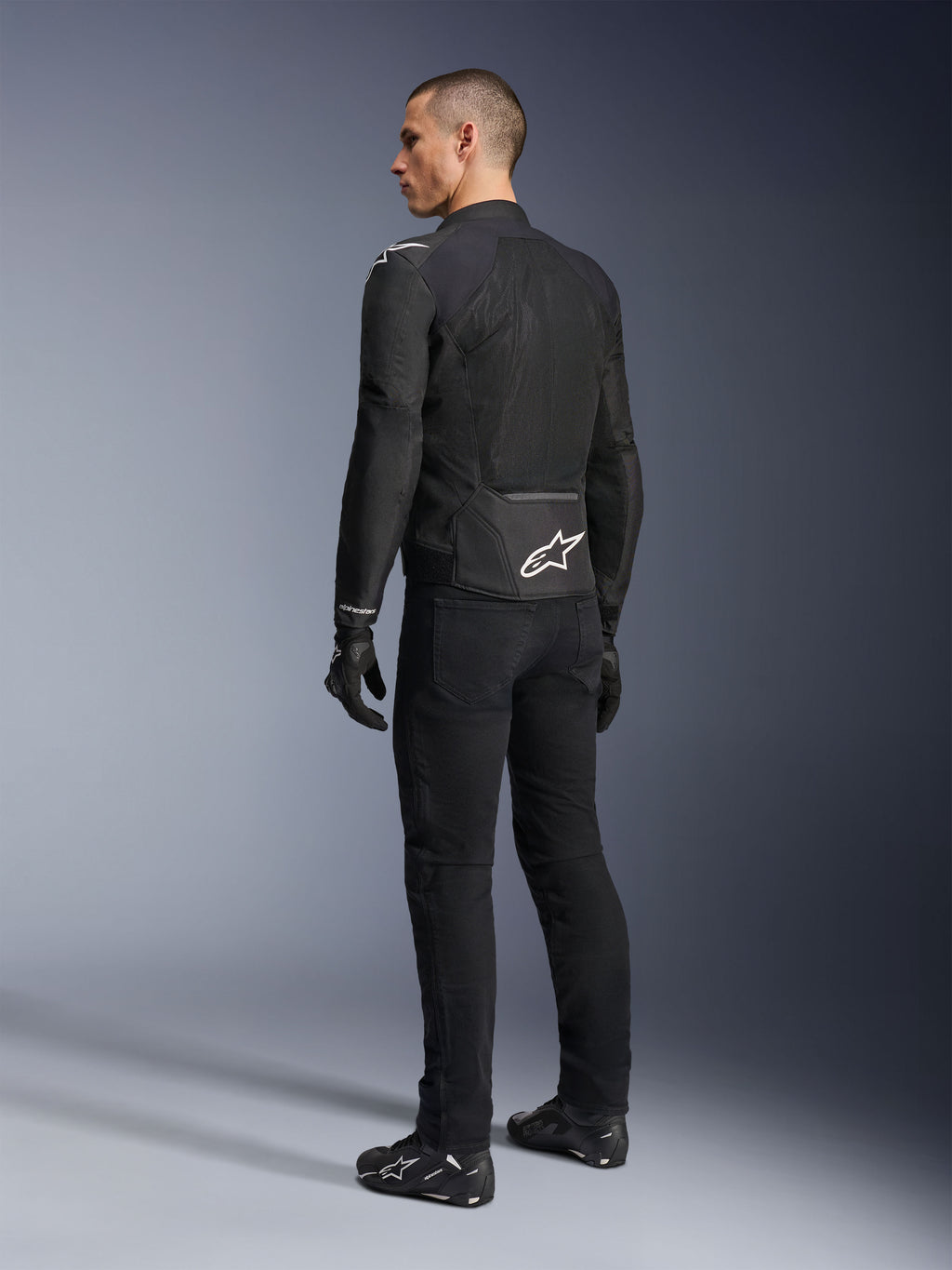 Alpinestars Viper Air V4 Jas, zwarte textiele motorjas, achteraanzicht op een mannelijk model, voorzien van grote tech mesh-panelen voor luchtstroom en een wit sterlogo op de onderrug, gedragen met bijpassende zwarte motorbroek en handschoenen