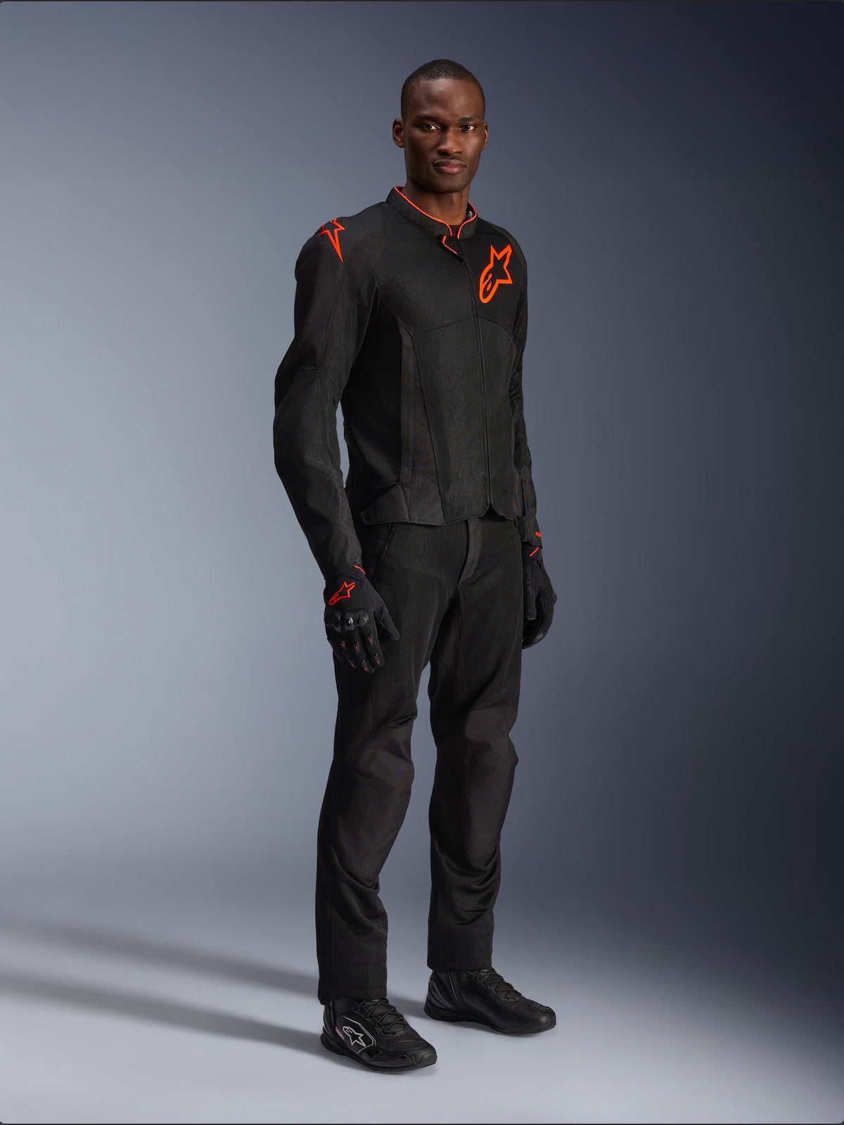 Model draagt Alpinestars Viper Air V4-jas in Black Red Fluo, voorzien van mesh panelen en rode sterlogo's op de borst en schouder, gecombineerd met bijpassende zwarte motorbroek, handschoenen en motorschoenen, professionele motorrijuitrusting voor op de weg