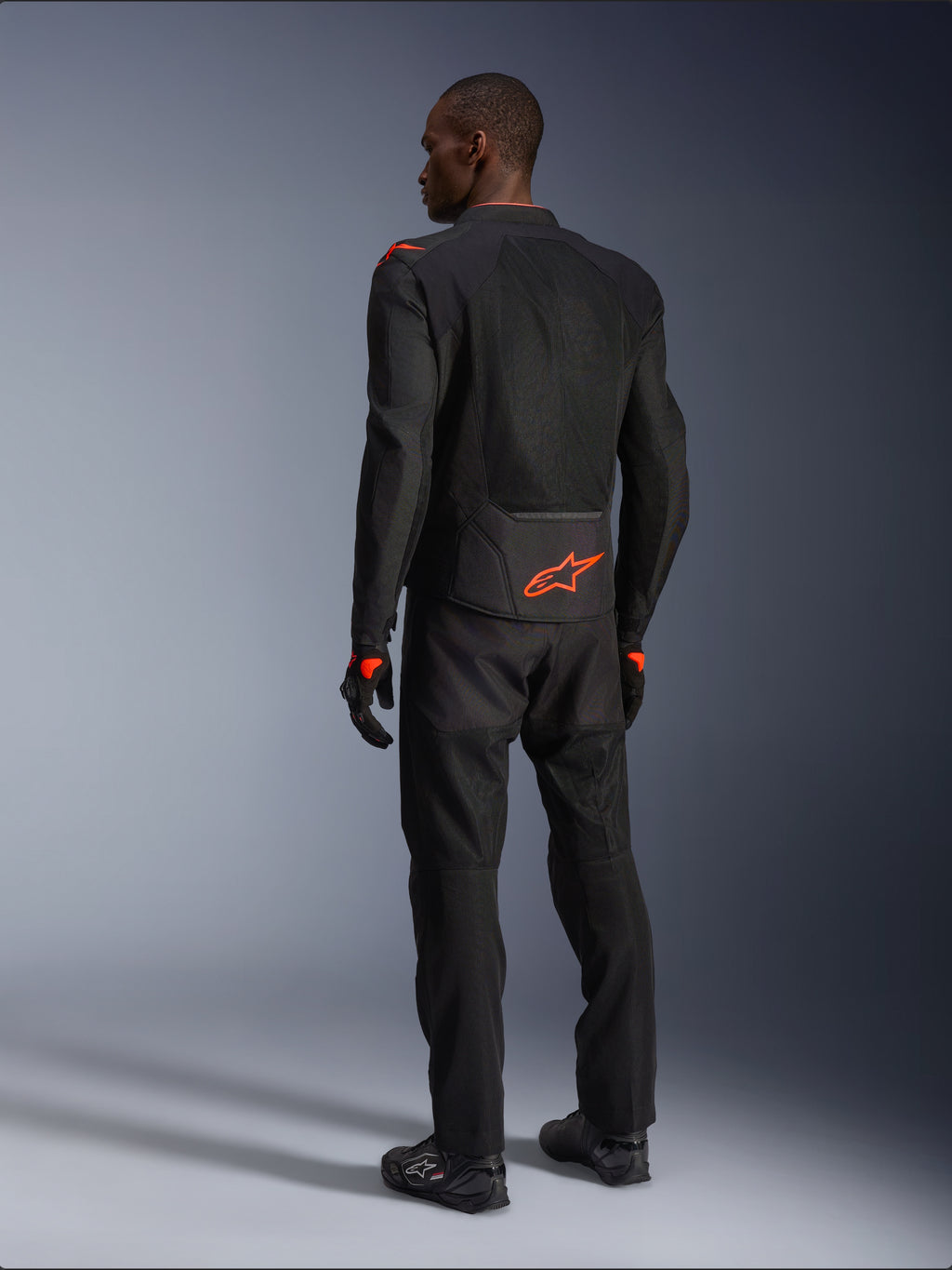 Mannelijke motorrijder van achteren gezien in de Alpinestars Viper Air V4 Jas, Black Red Fluo, met een oranje Astars-logo op de onderrug en schouders, mesh-textielconstructie, gecombineerd met bijpassende zwarte motorbroek en laarzen.
