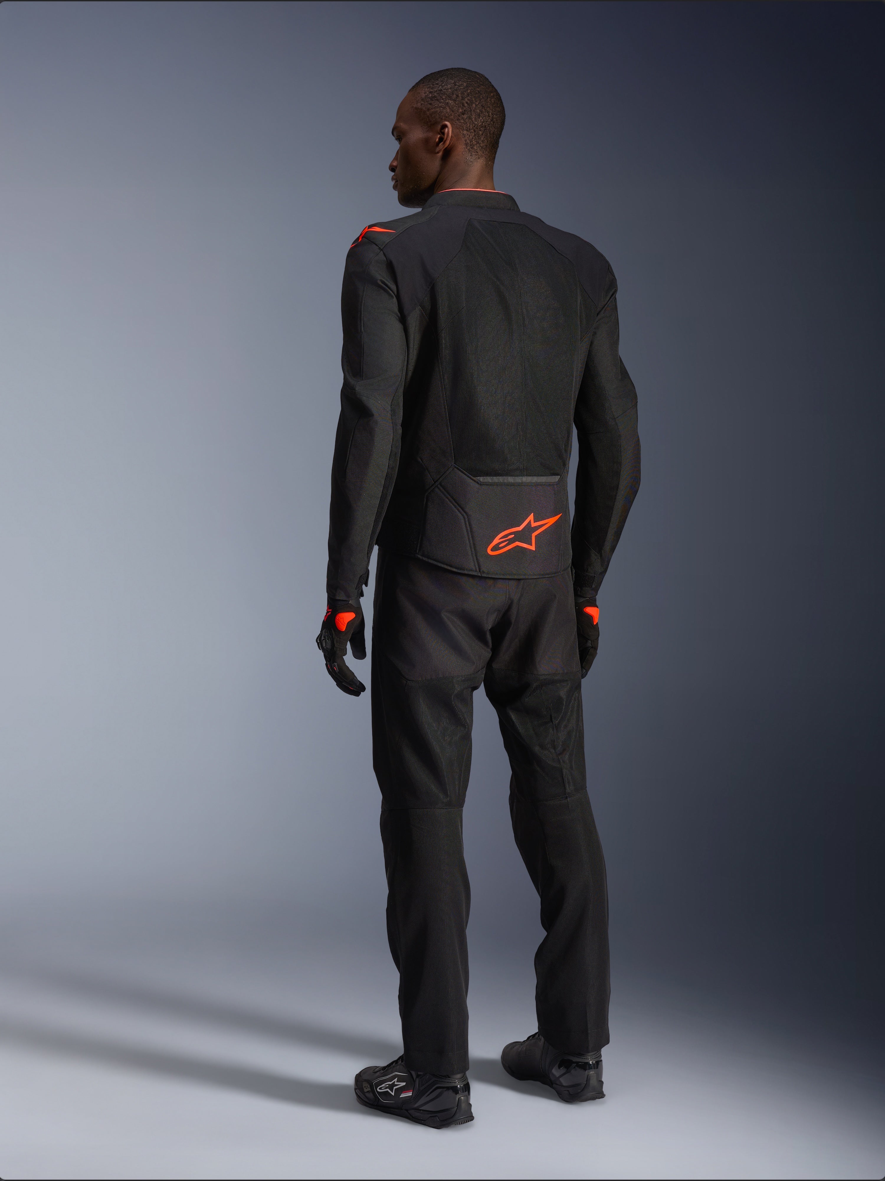 Mannelijke motorrijder van achteren gezien in de Alpinestars Viper Air V4 Jas, Black Red Fluo, met een oranje Astars-logo op de onderrug en schouders, mesh-textielconstructie, gecombineerd met bijpassende zwarte motorbroek en laarzen.
