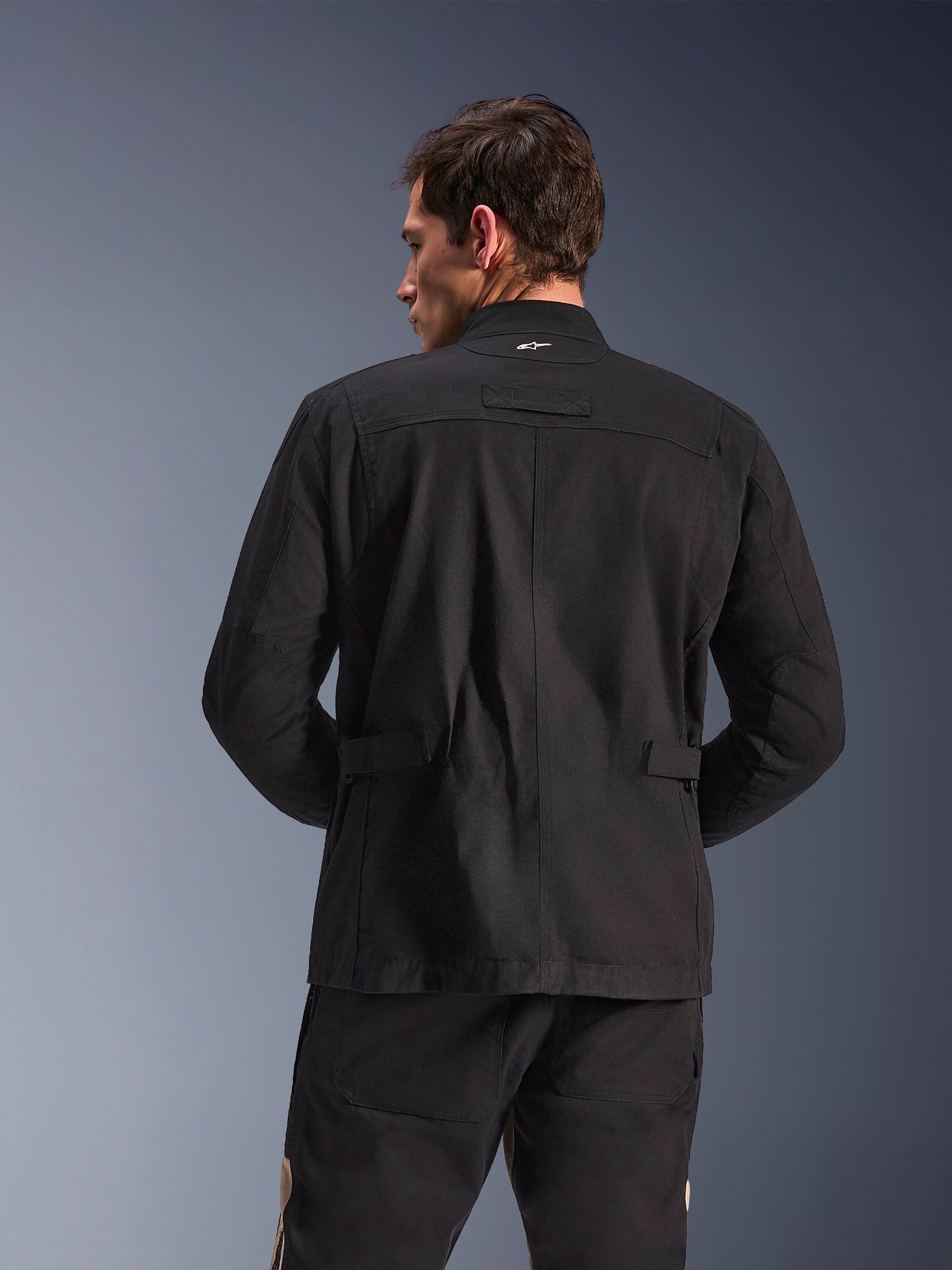 Alpinestars Dice Jacket, urban motorjas, zwart, achteraanzicht met geplooide schouders en verstelbare taillebandjes, veldjas-esthetiek met klein wit logo op de kraag, beschermende textielkleding voor stadsritten