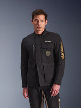 Alpinestars Dice Jacket, zwarte motorjas, gedragen door model, voorzien van gouden Alpinestars-logo's, meerdere functionele zakken, ronde spiraalvormige patch en urban field jacket-stijl