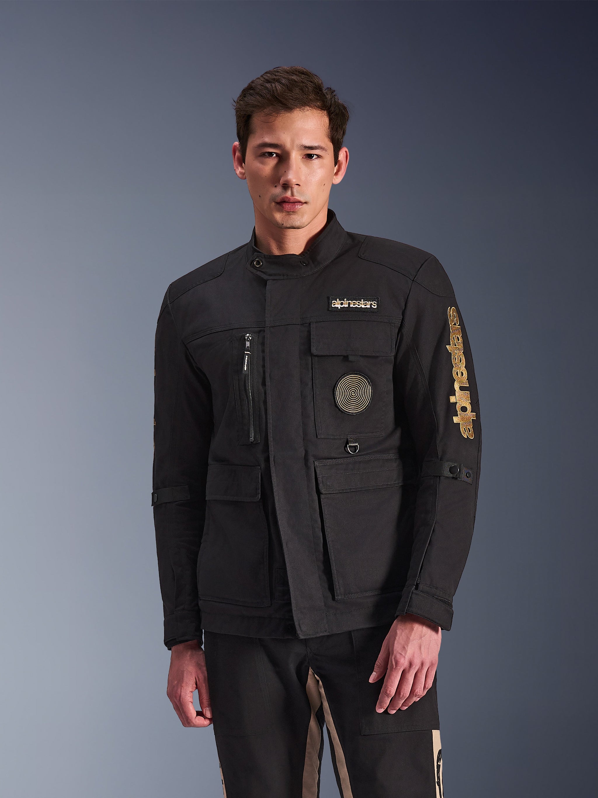 Alpinestars Dice Jacket, zwarte motorjas, gedragen door model, voorzien van gouden Alpinestars-logo's, meerdere functionele zakken, ronde spiraalvormige patch en urban field jacket-stijl