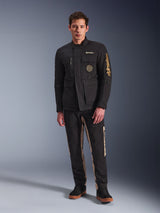 Mannelijk model draagt de Alpinestars Dice Jacket, zwarte urban motorjas met vintage veldjas-stijl, vier functionele voorzakken, Alpinestars-branding op de mouw en borst, gecombineerd met een zwart-beige broek, moderne stedelijke motoruitrusting