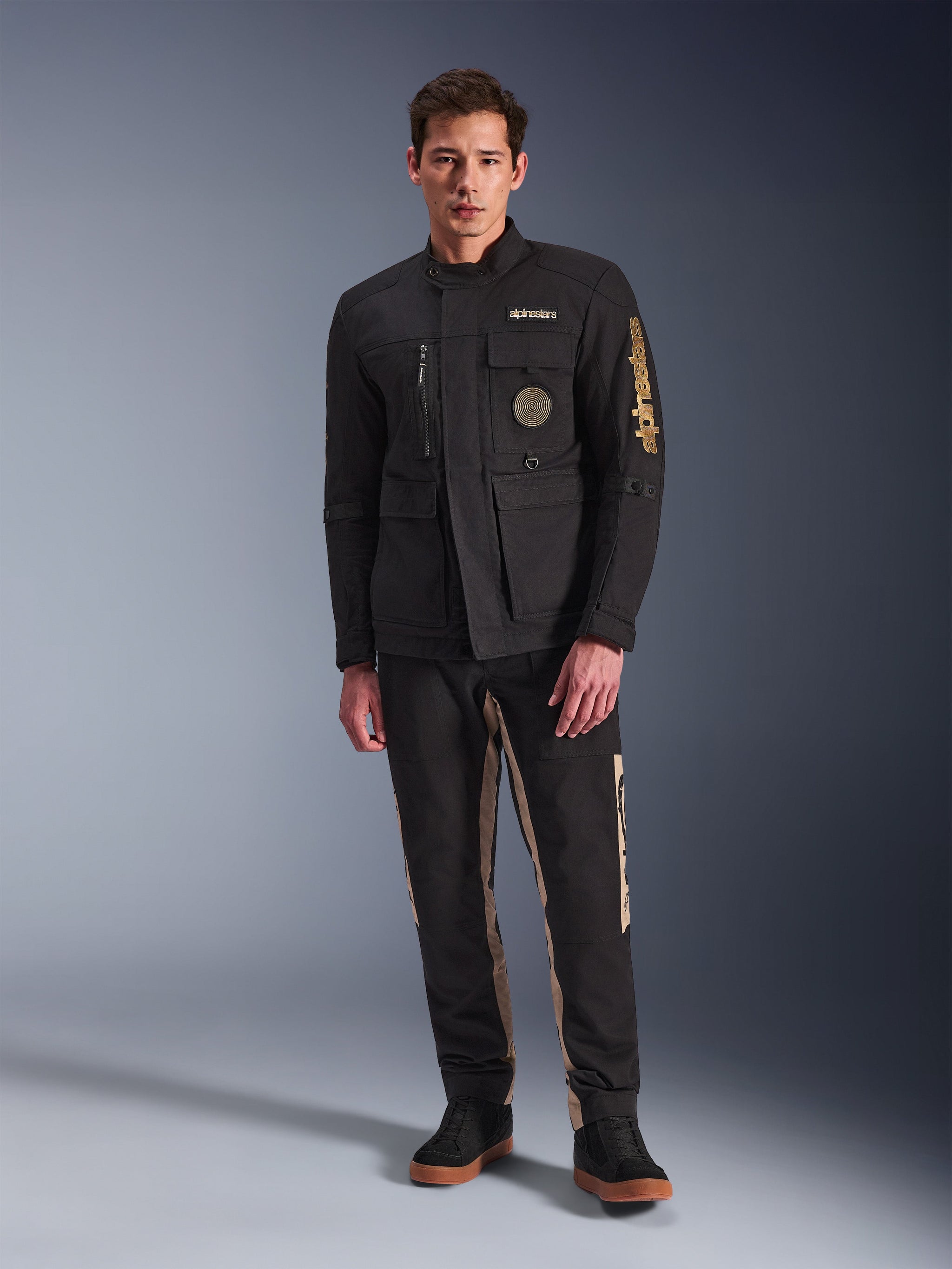 Mannelijk model draagt de Alpinestars Dice Jacket, zwarte urban motorjas met vintage veldjas-stijl, vier functionele voorzakken, Alpinestars-branding op de mouw en borst, gecombineerd met een zwart-beige broek, moderne stedelijke motoruitrusting