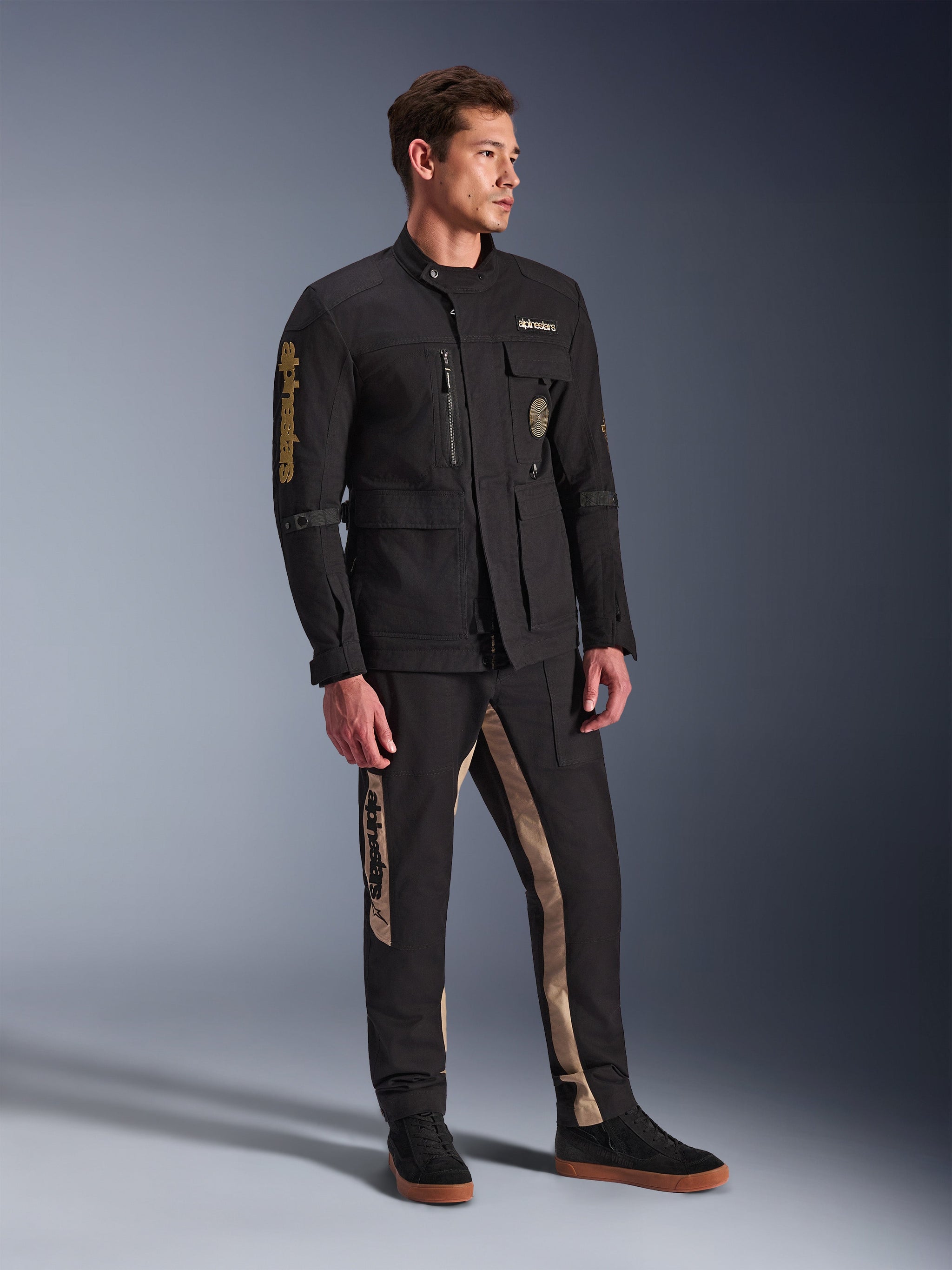 Mannelijk model in Alpinestars Dice Jacket in het zwart, urban motorjas met vintage veldjas-esthetiek, voorzien van meerdere functionele zakken en gouden branding op de mouw, gecombineerd met bijpassende zwart-beige broek en zwarte sneakers