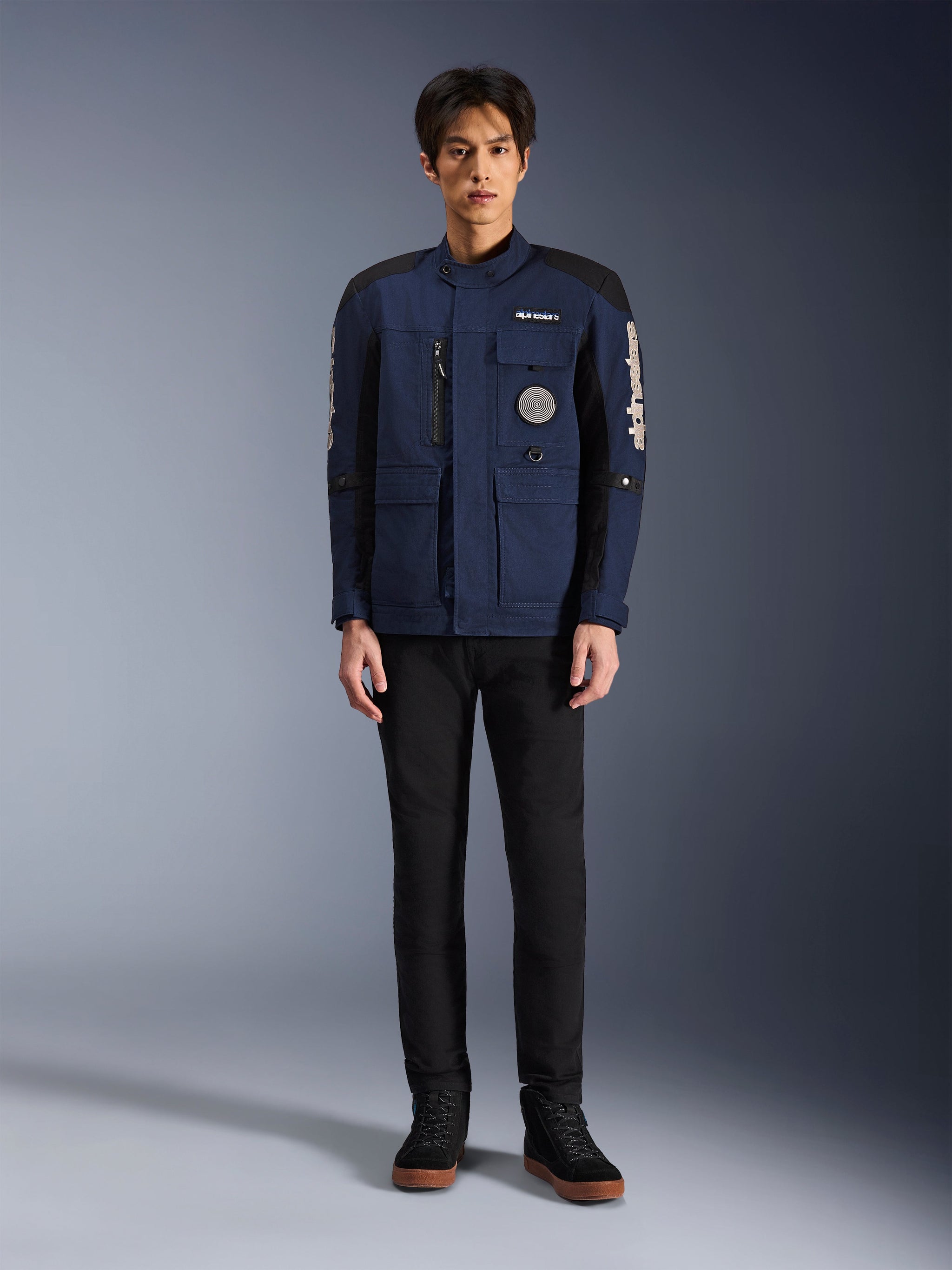 Alpinestars Dice Jacket, urban motorjas, donkerblauw en zwart, gedragen door een model met zwarte broek en zwarte schoenen, modern veldjas-ontwerp met meerdere zakken, verticale borstritssluiting en witte Alpinestars-branding op de mouwen.